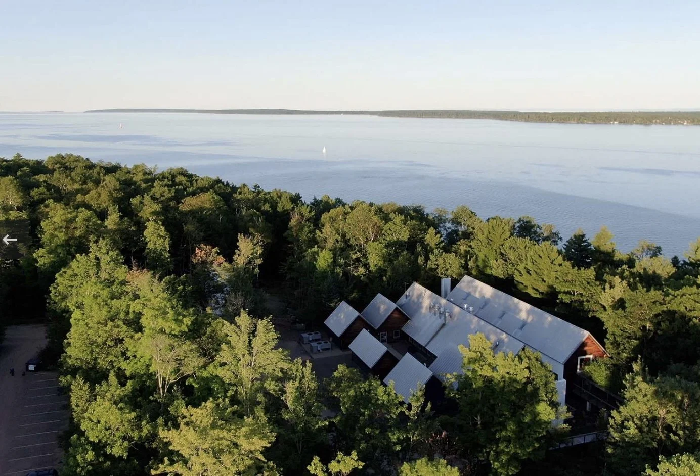 Aerial view of a lakeside area with a forest, buildings with metal roofs, and a large body of water in the background.
