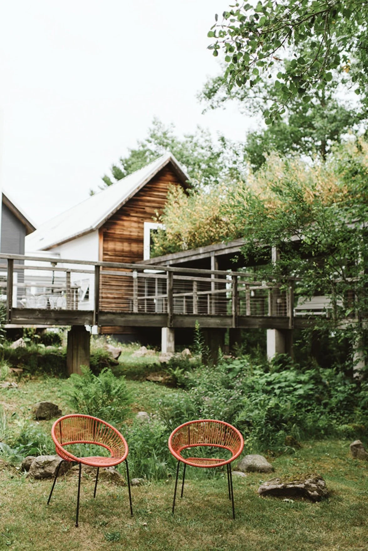 Two red outdoor chairs on a grassy yard with rock decorations, greenery, trees, and a wooden house with a deck in the background.