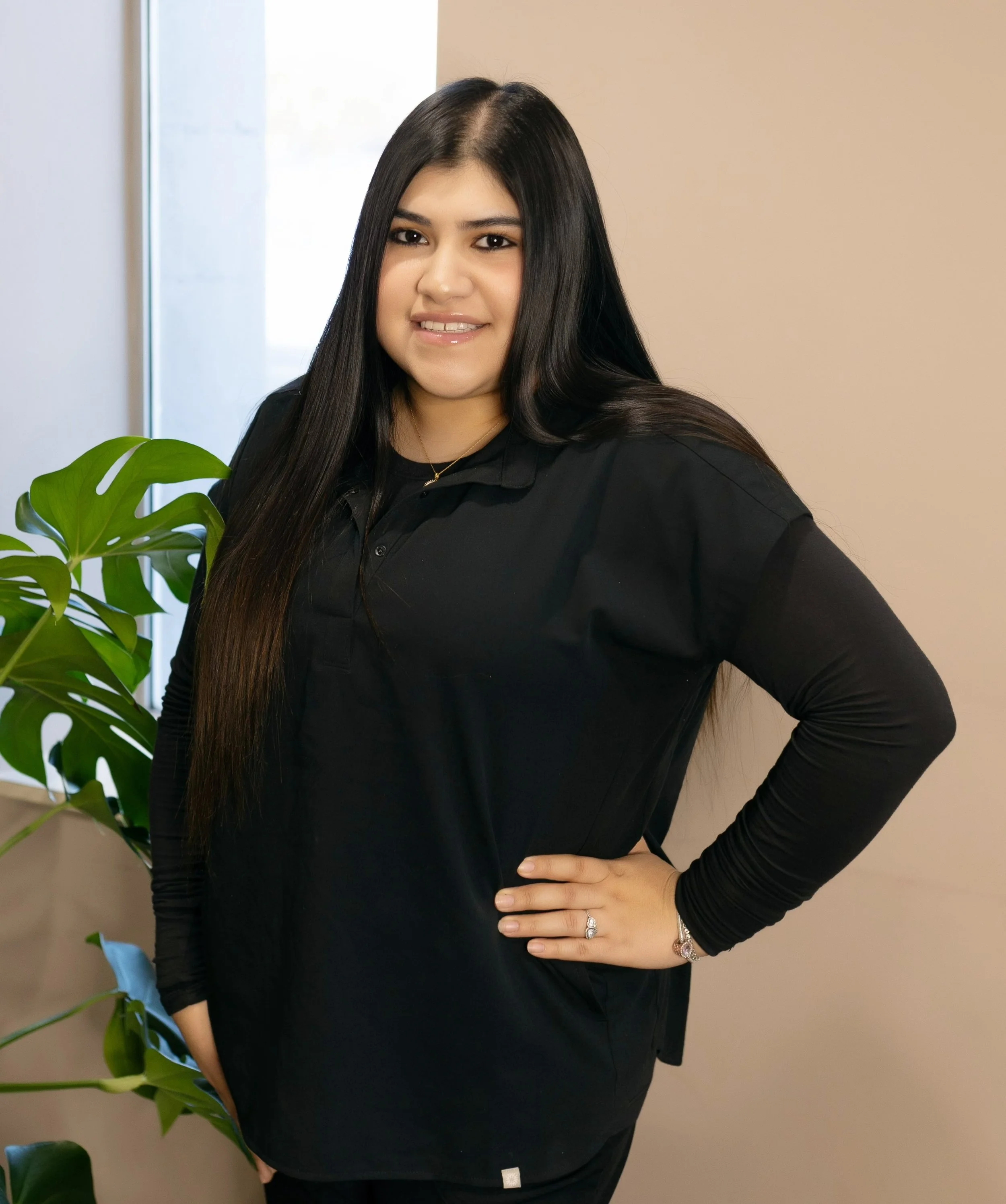 A woman with long dark hair, wearing a black long-sleeve shirt, standing indoors next to a green plant, with her hand on her hip, smiling at the camera.