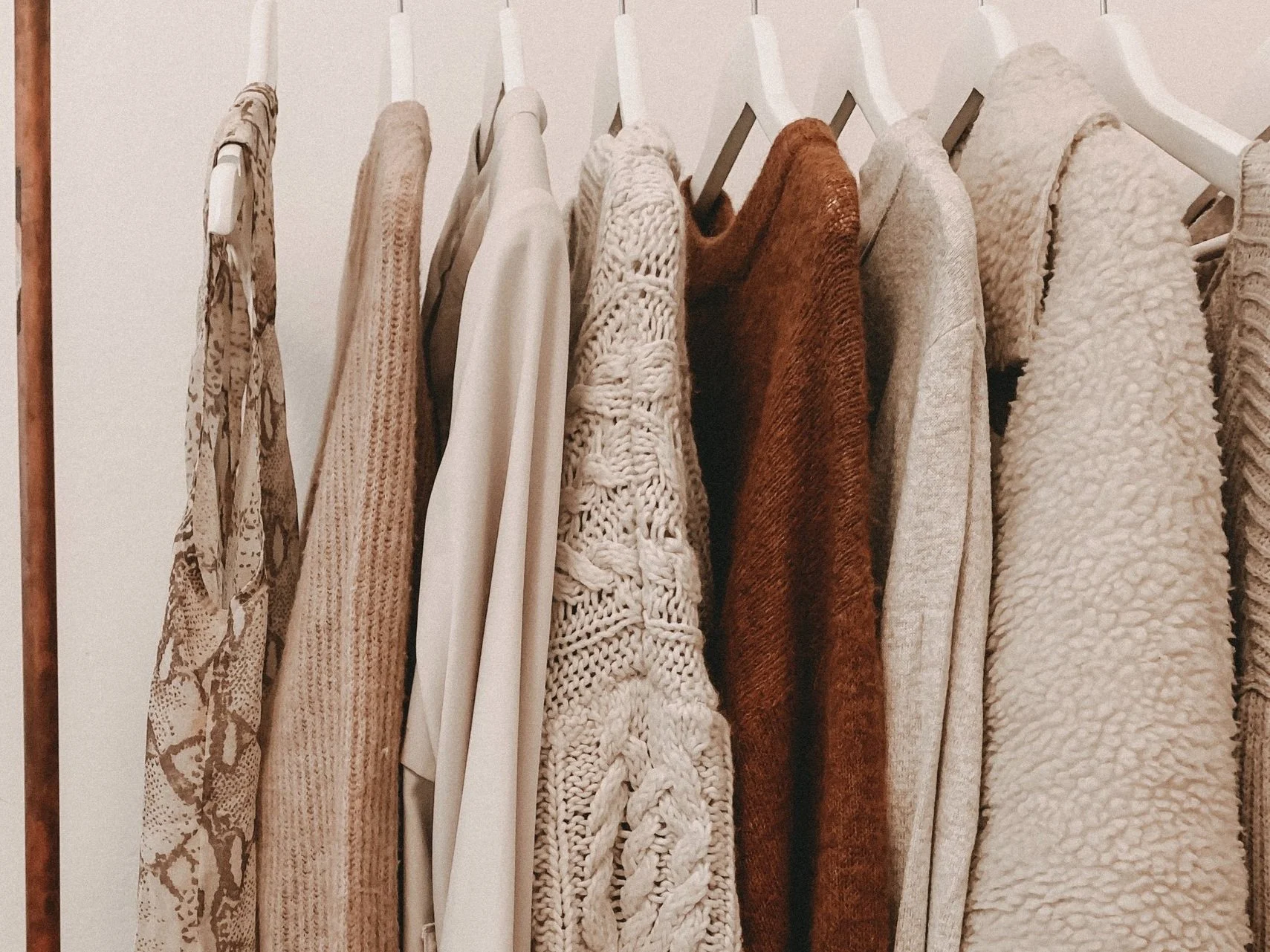 Assorted neutral-toned sweaters and tops hanging on white hangers on a clothing rack.