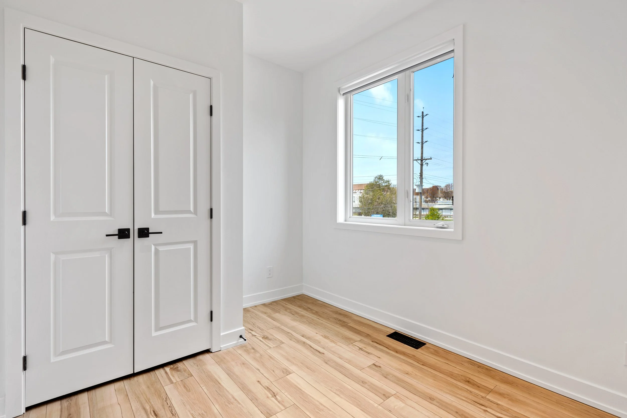 Empty room with light wooden floor, white walls, and a window with outside view, featuring a closed white double-door closet on the left.