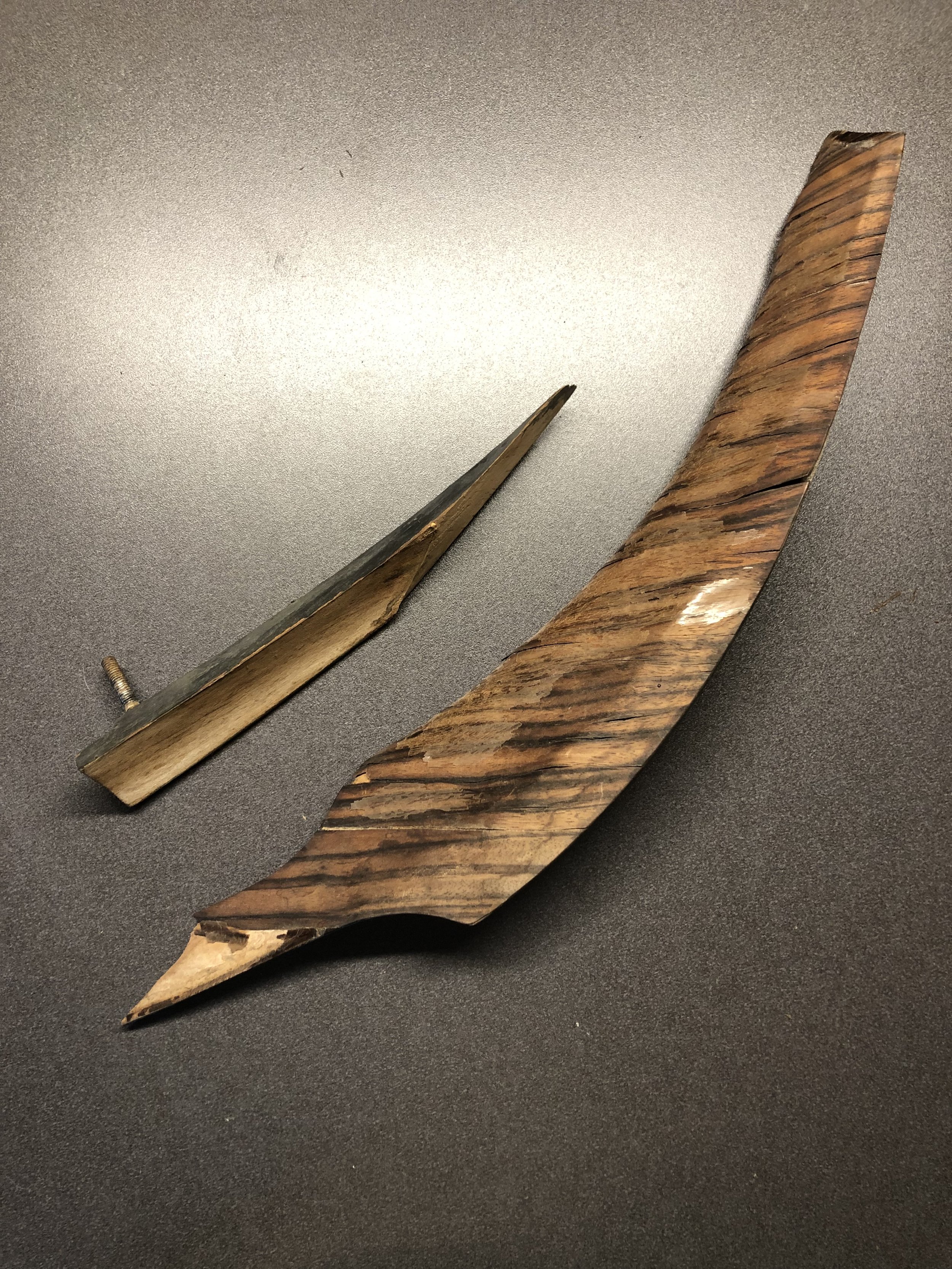 A foot-long piece of wood that is gently curved and also smoothly contoured on one face is broken in half forming a sharp point on each resulting piece. The two pieces sit on a gray background. A small, threaded metal stud protrudes from the back of