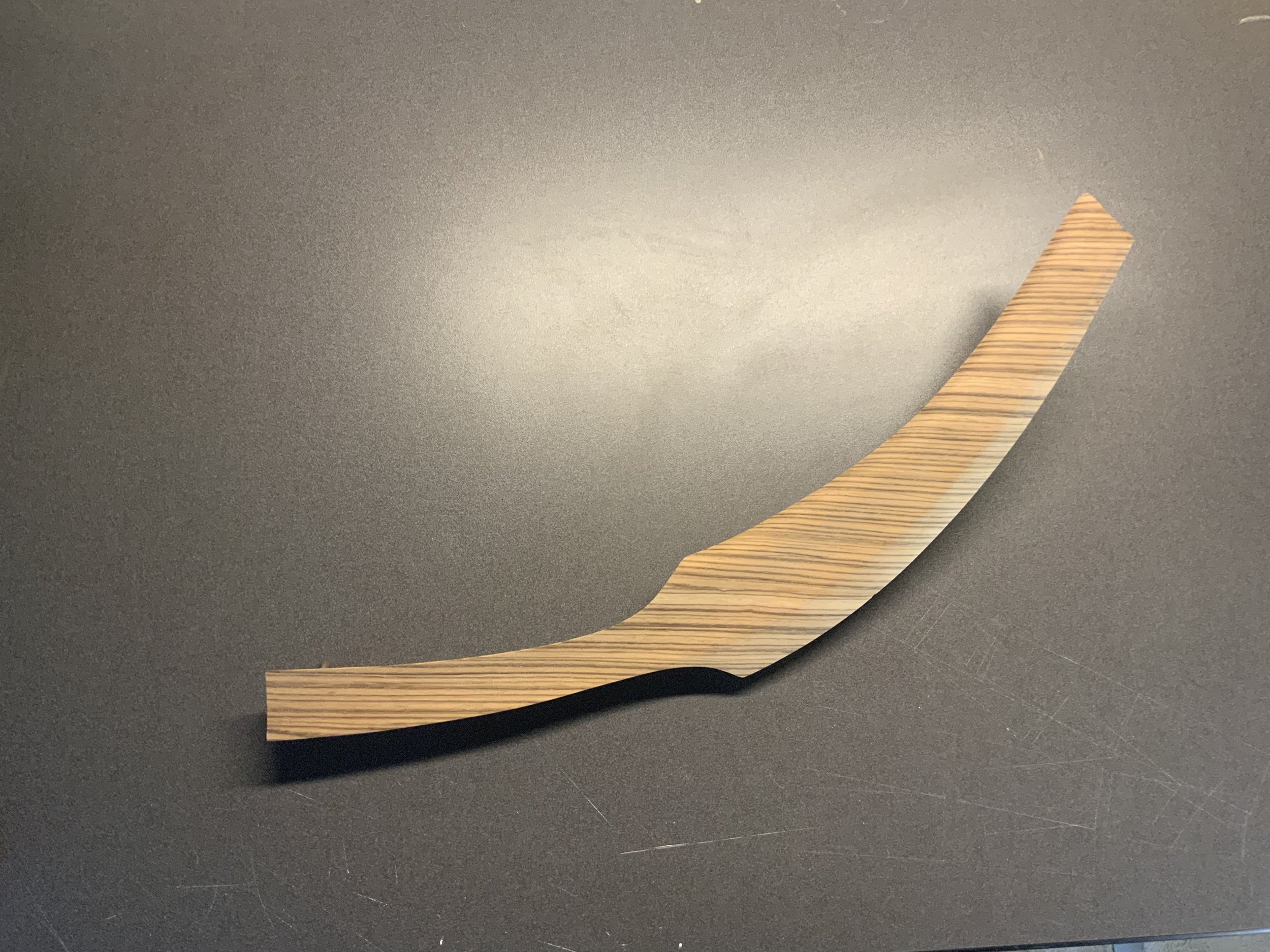 Sitting on a gray background and well-lit, a foot-long piece of wood that is gently curved and also smoothly contoured on one face is veneered in a wood grain composed of very straight stripes of alternating dark brown and medium brown tones.