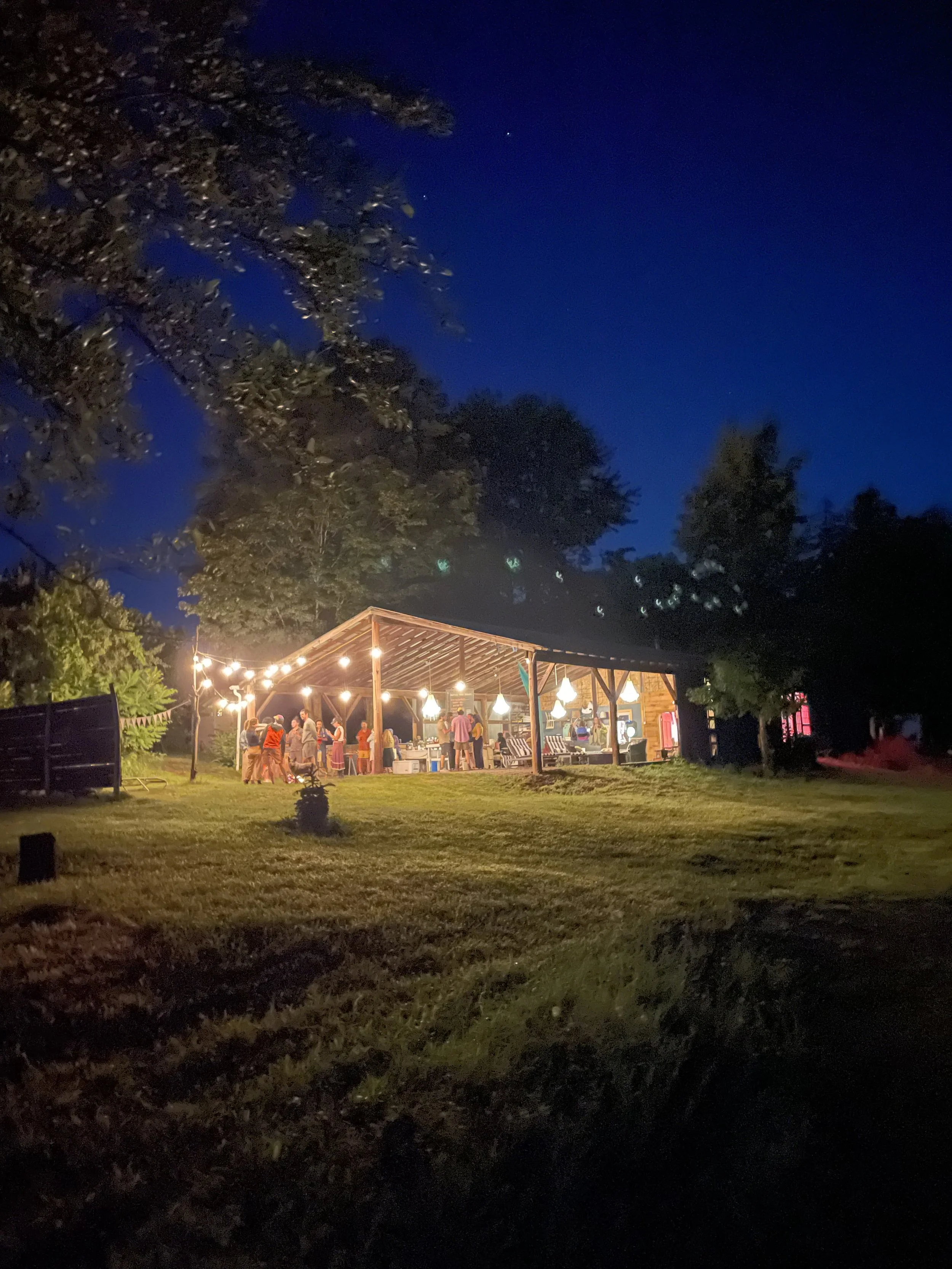 Outdoor covered barn venue space with string lights, chandeliers, people gathering around a fire, dark night sky and brightly lit interior.