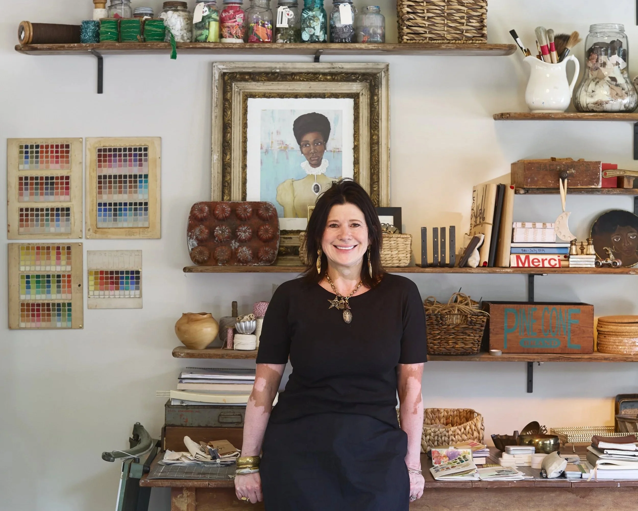 A woman with dark hair wearing a black dress, smiling, standing in front of a cluttered desk with art supplies and books. Behind her is a wall with shelves holding jars, baskets, and framed artwork, including a portrait of a woman with an afro hairstyle.