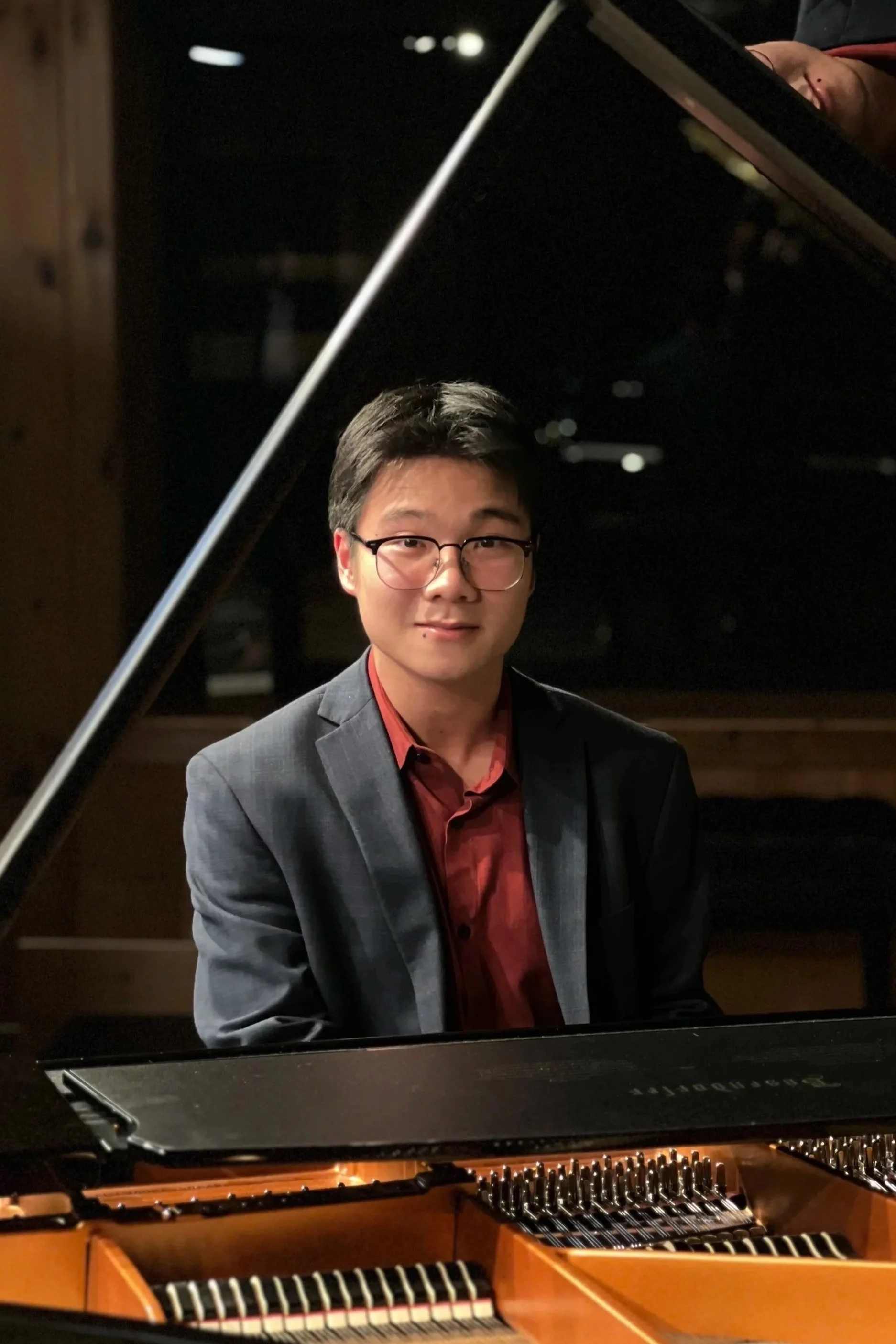 A man wearing glasses and a business suit sitting at a grand piano, looking at the camera indoors in a dimly lit setting.