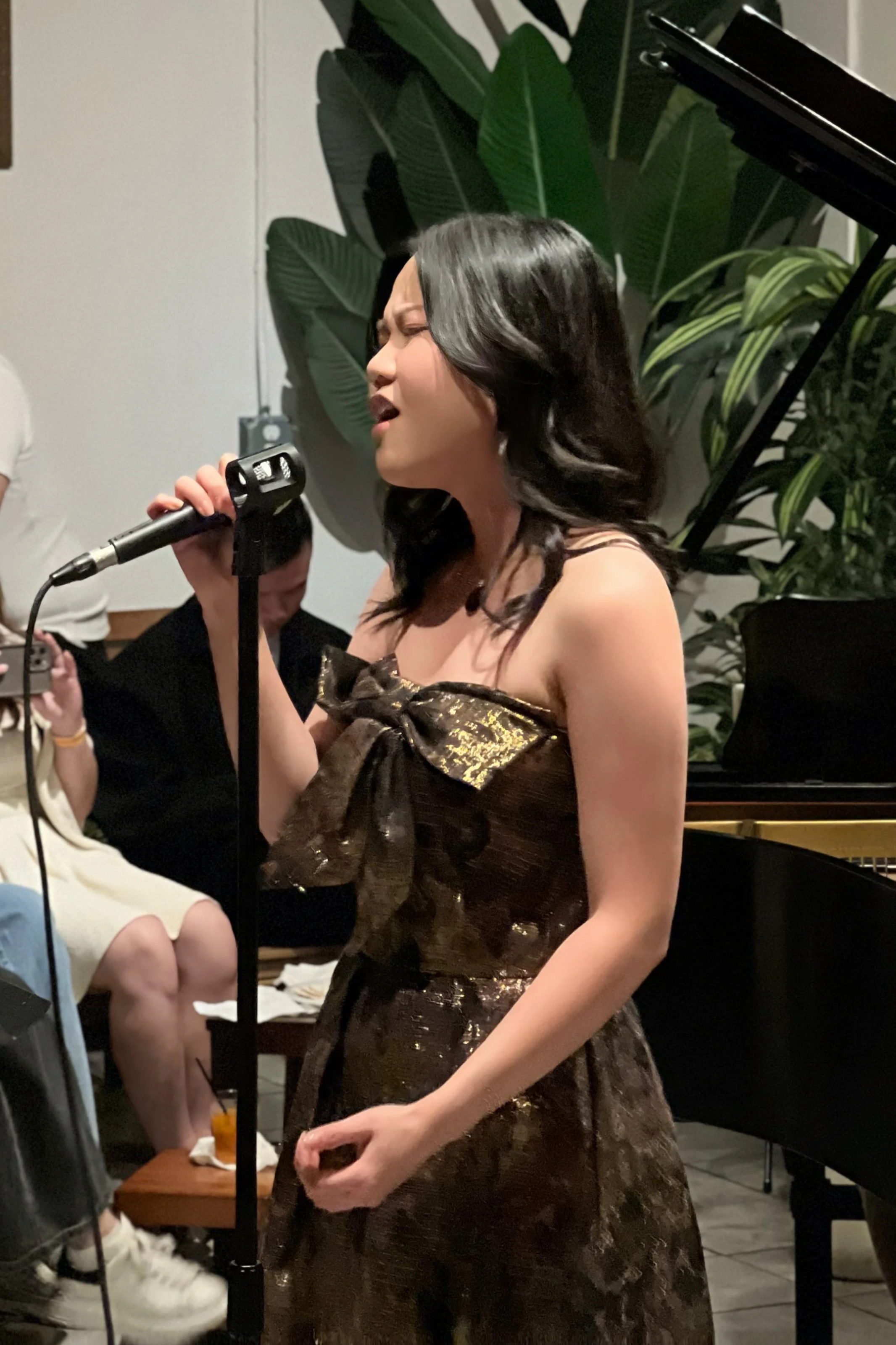 A woman with black hair singing into a microphone at a live music performance, wearing a dark, shiny dress with a bow detail, next to a grand piano, in a setting with large green plants.
