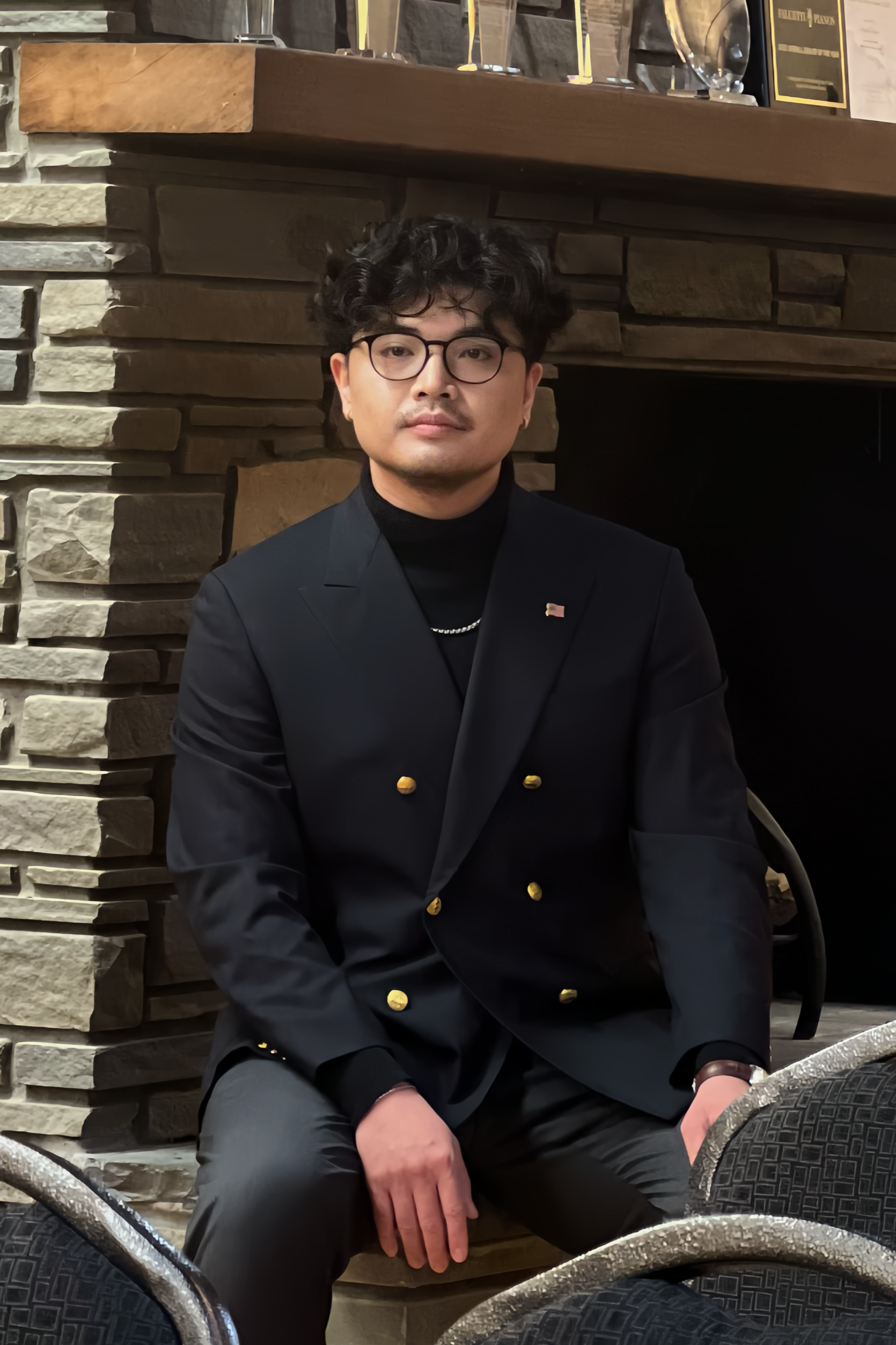 A man with glasses and curly hair sitting indoors in front of a stone fireplace, wearing a black blazer with gold buttons and a small American flag pin.