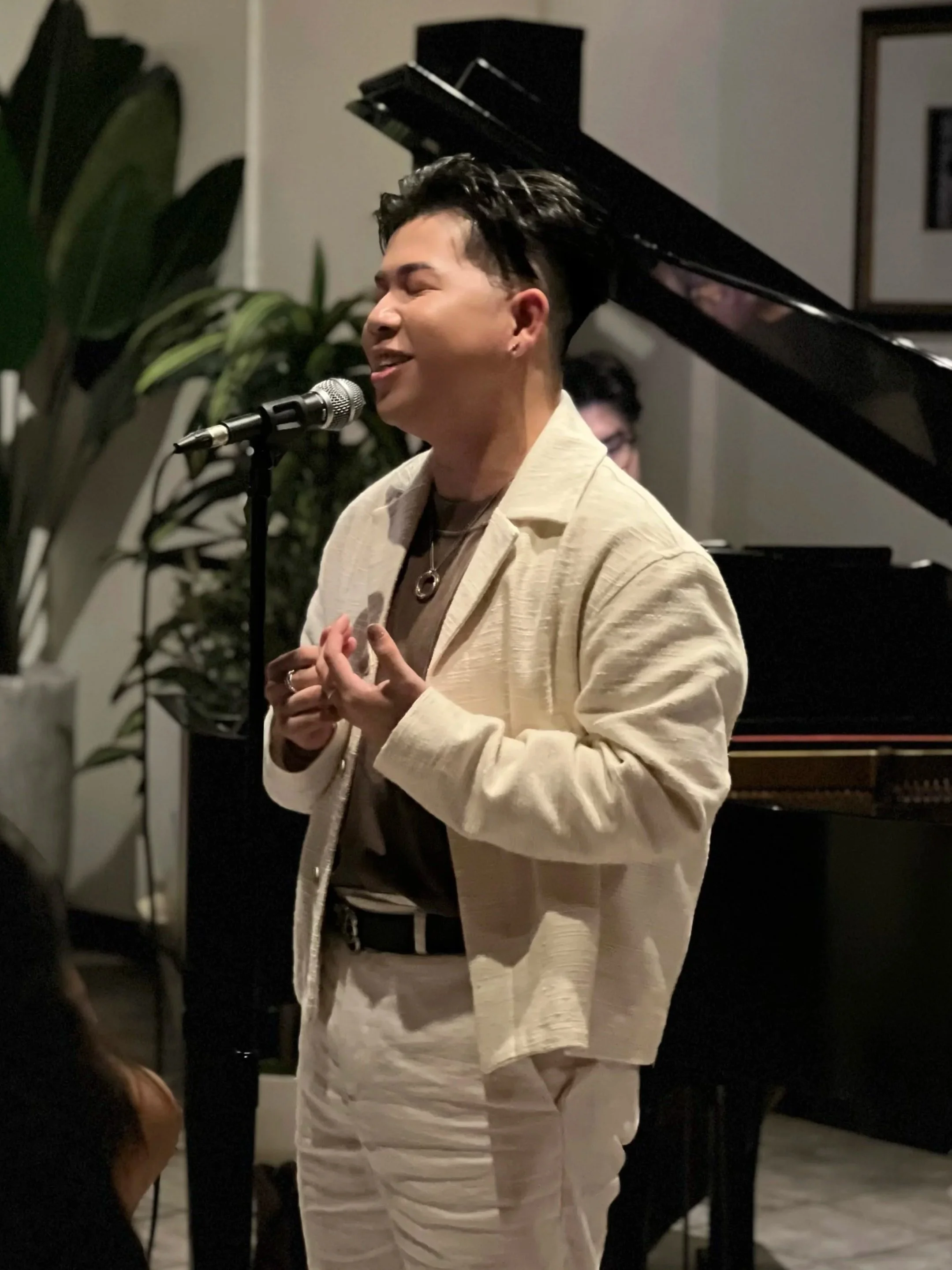A person singing into a microphone with eyes closed, standing beside a grand piano in a cozy, dimly-lit room.