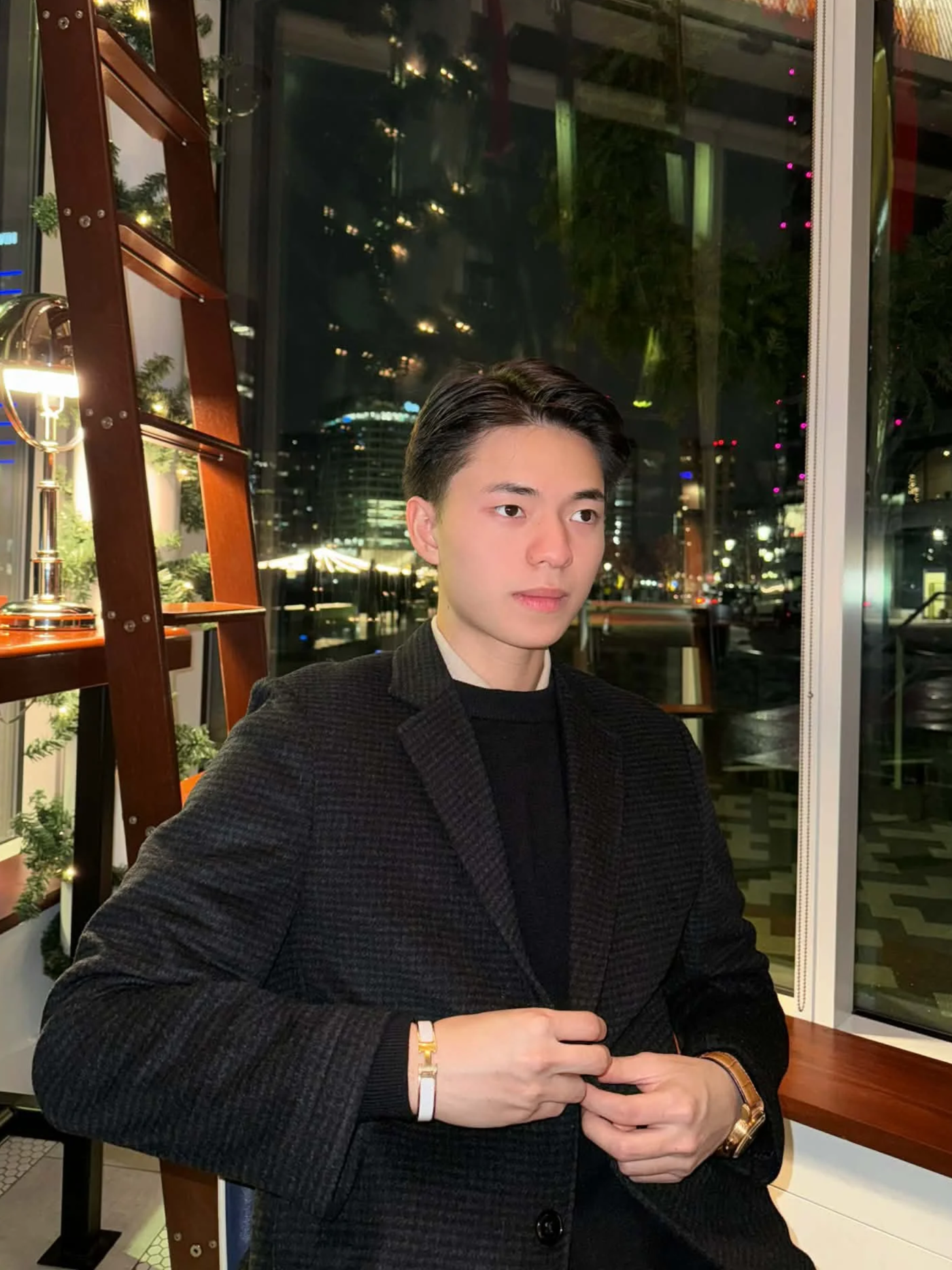 Young man with dark hair wearing a black blazer and watch, sitting in a modern indoor space at night with city lights visible through large windows.