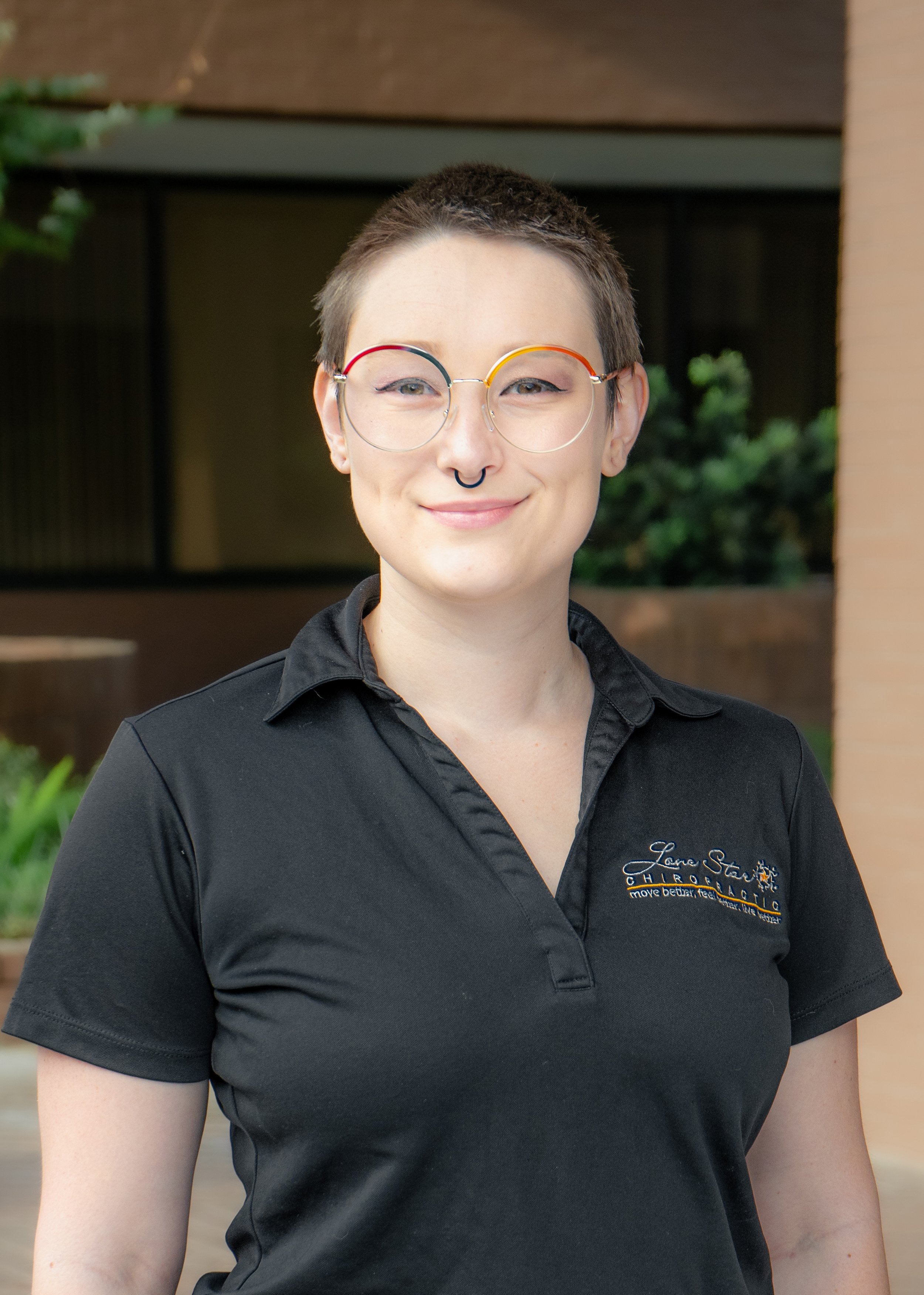 Person with short hair, wearing round glasses with colorful frames and a black polo shirt, standing outside.