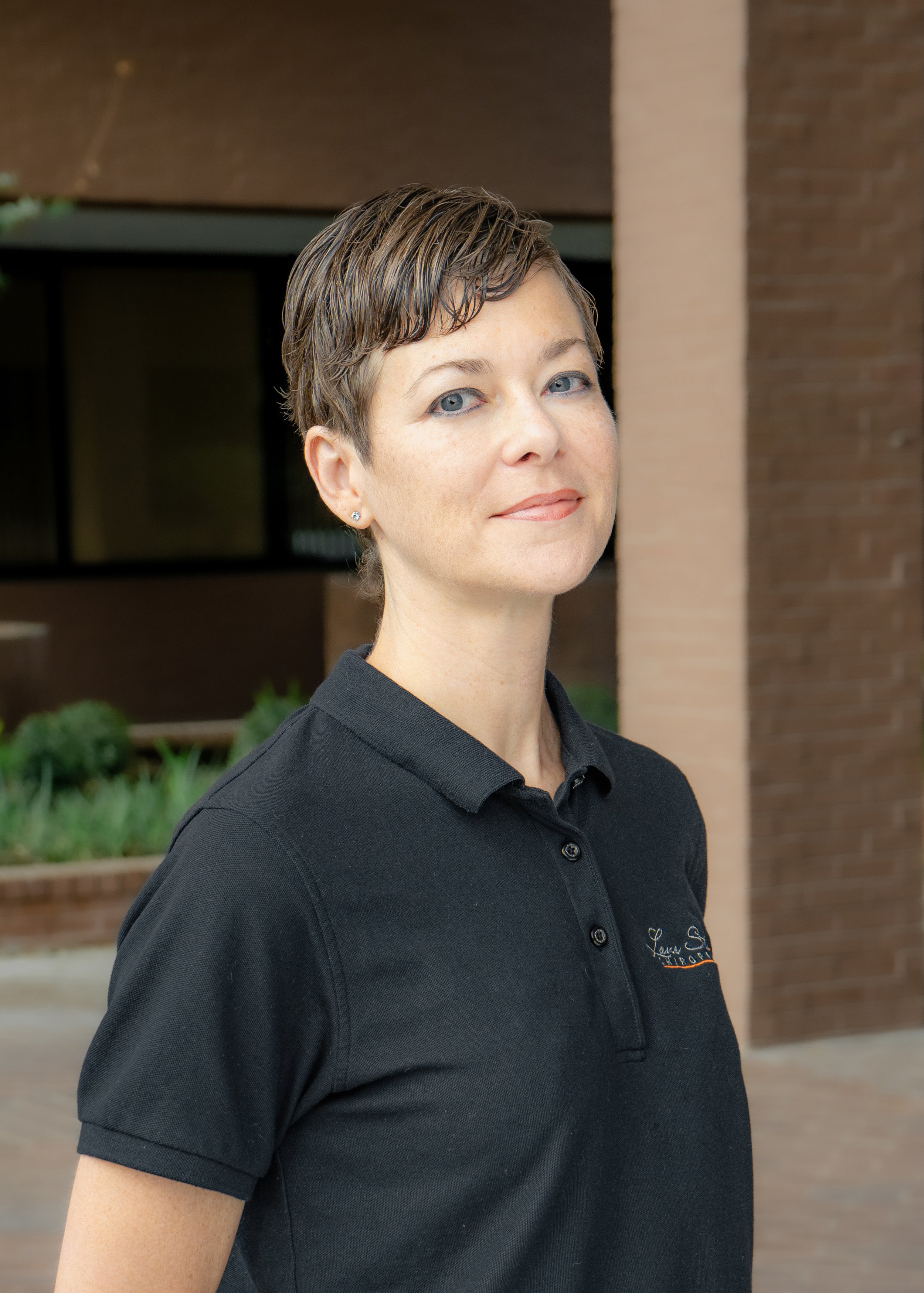 Person with short hair wearing a black polo shirt, standing outside near a brick building.