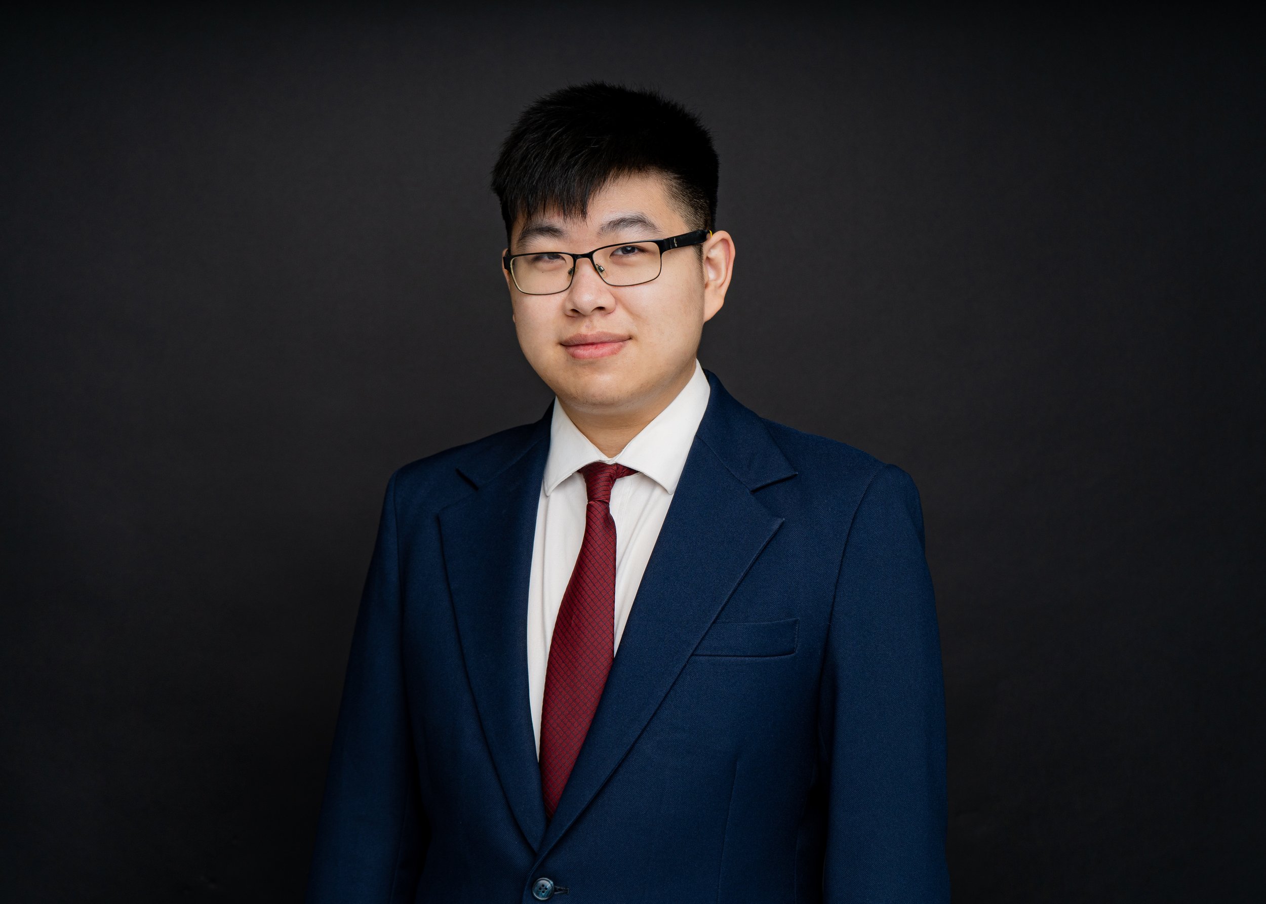 Person wearing glasses and a suit with a red tie, standing against a black background. Clutch City Insurance Staff Headshot.