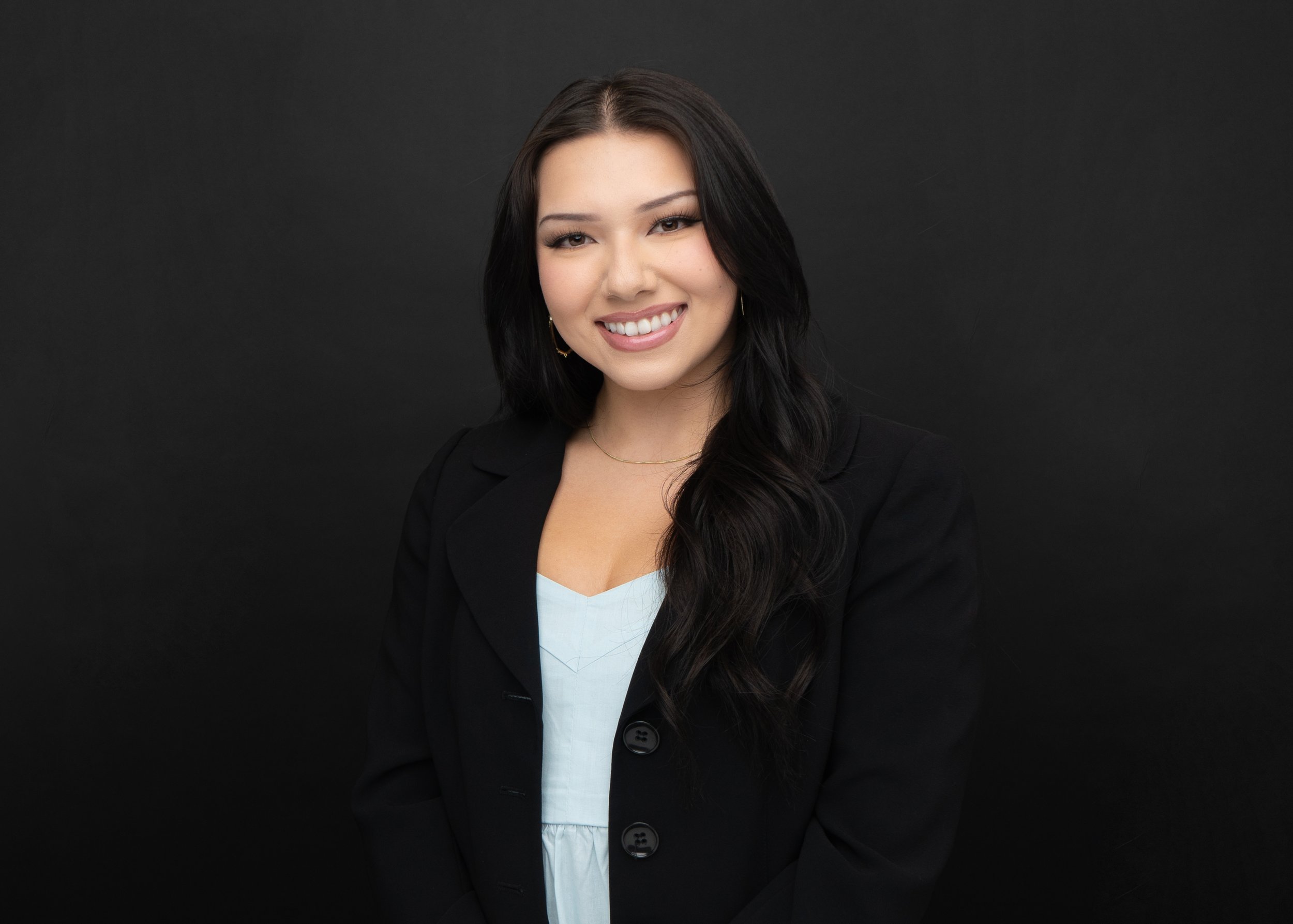 Smiling woman with long dark hair, wearing a black blazer and light blue top, against a black background. Clutch City Insurance Staff Headshot.