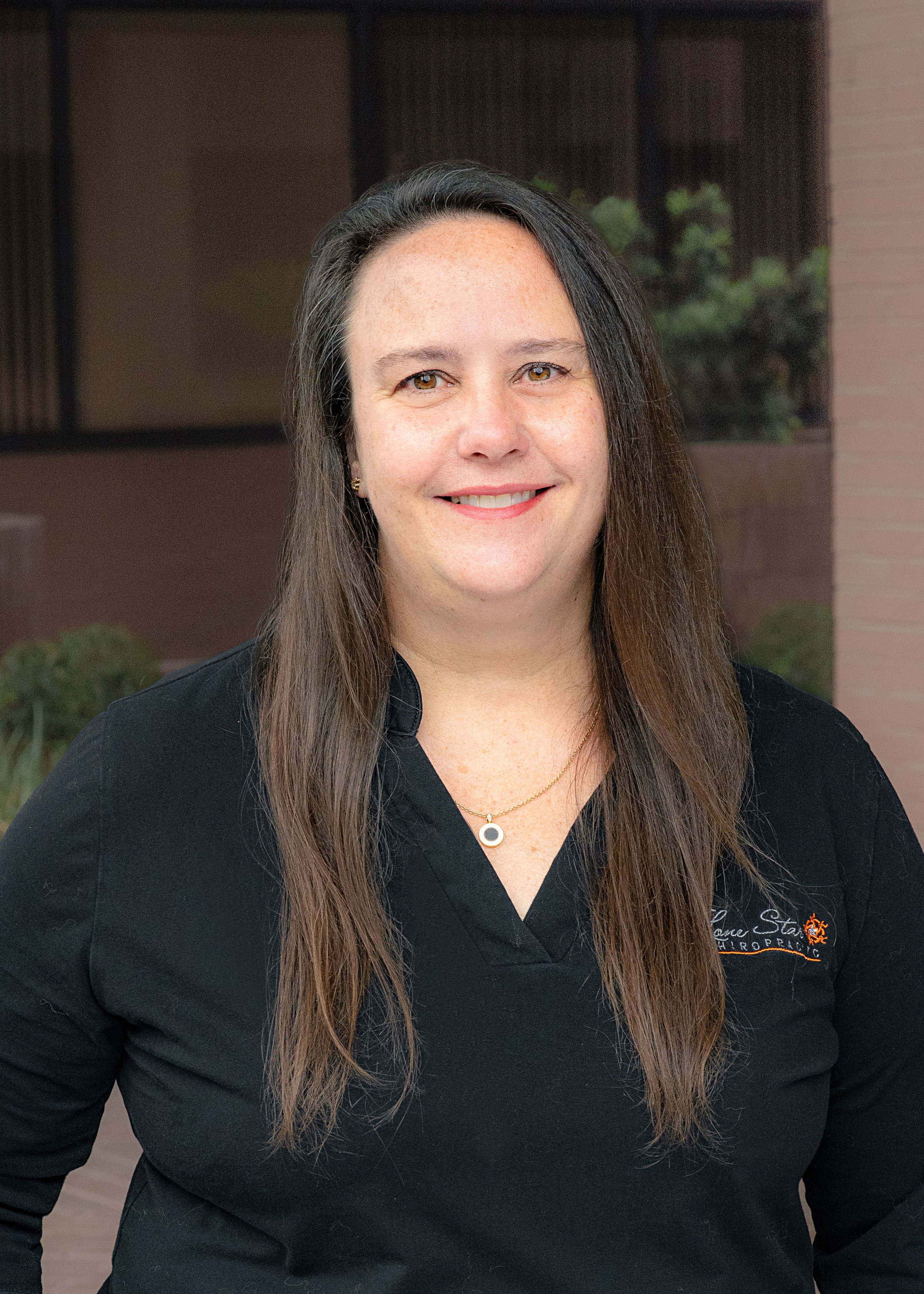 Smiling woman with long hair wearing a black shirt and necklace standing in front of a building.