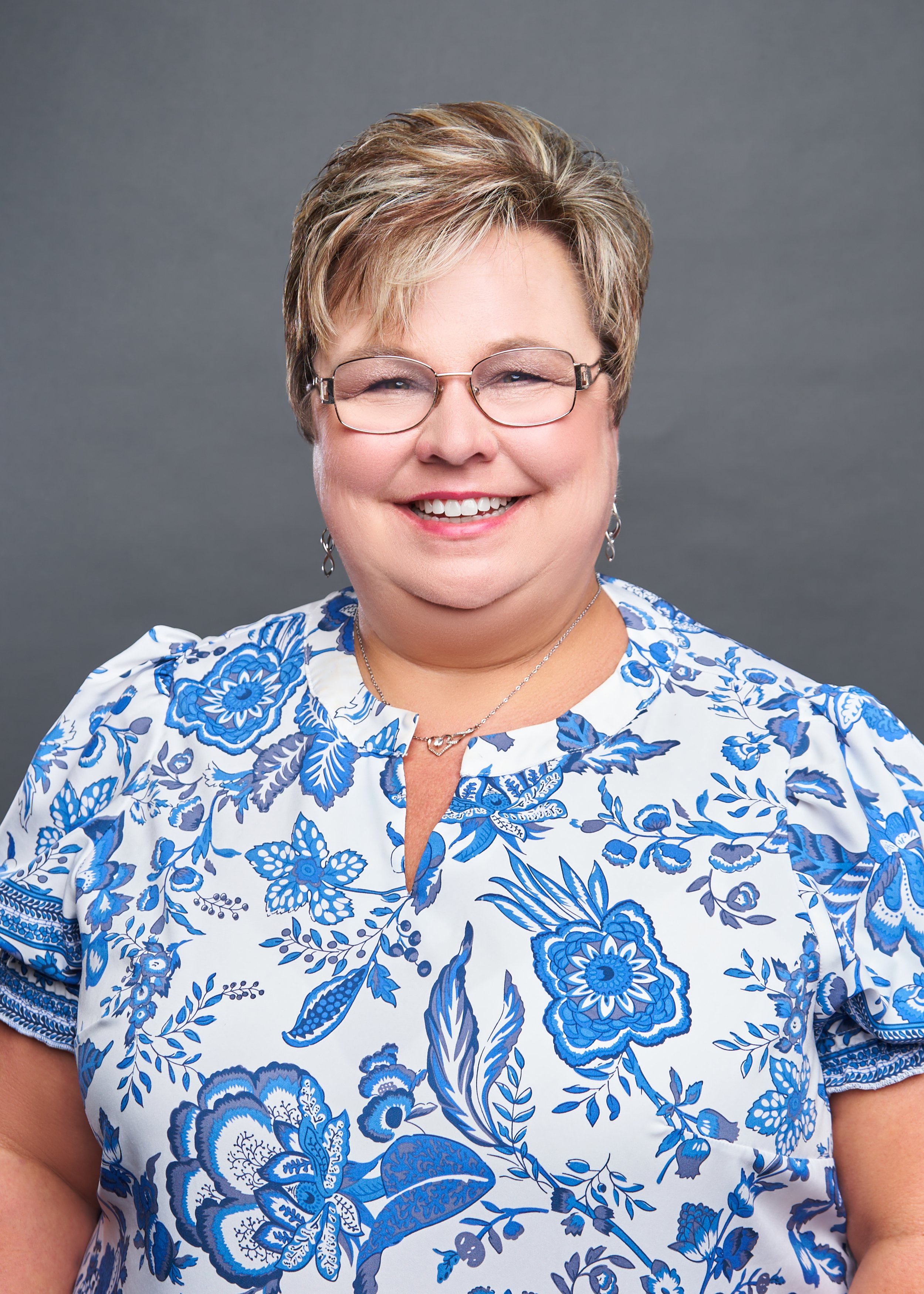 Smiling woman with short hair, glasses, wearing a blue floral dress, against a grey background.