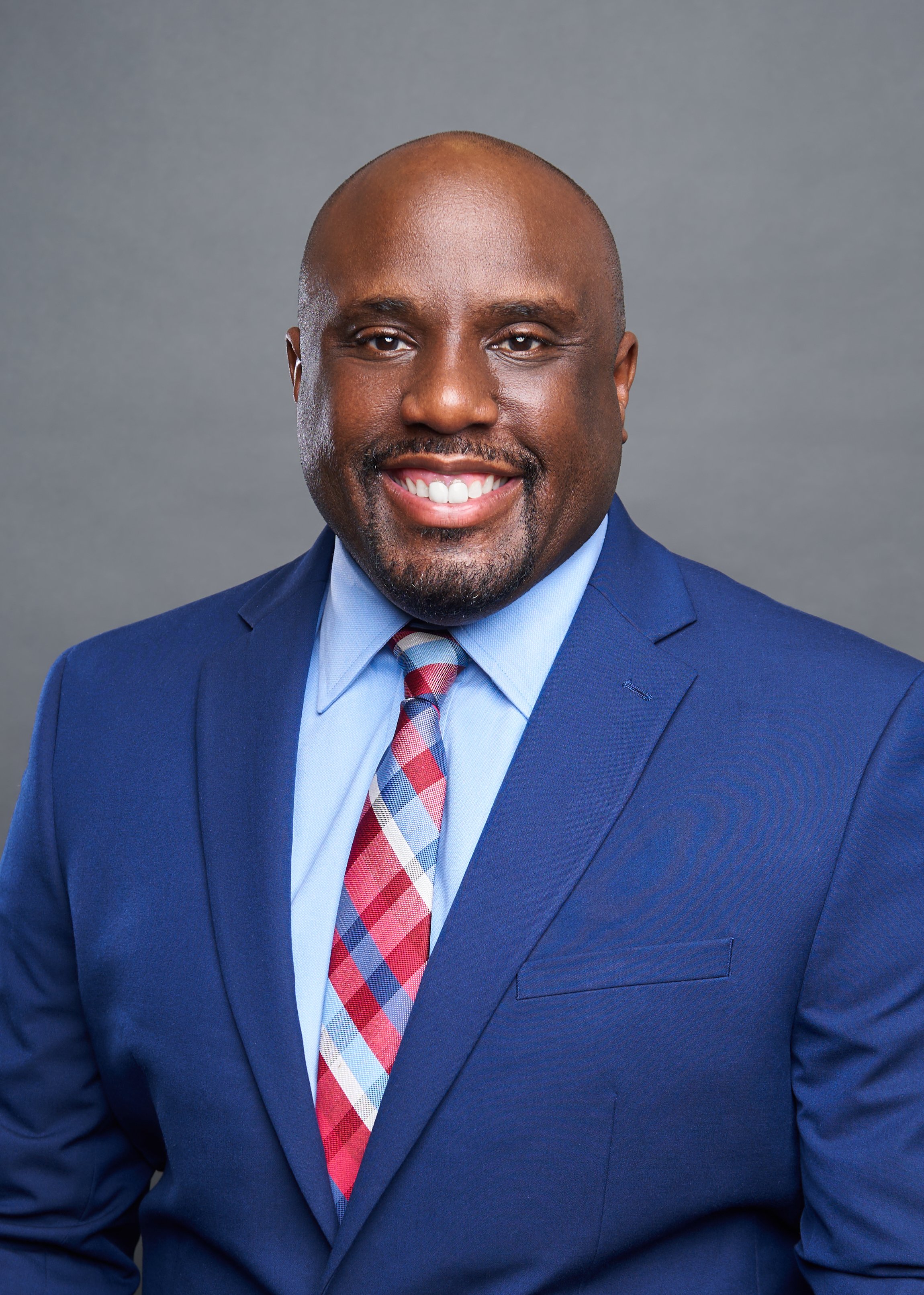 Smiling man in a blue suit and multicolored tie against a gray background.