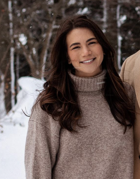 A young woman smiling outdoors in winter, wearing a beige turtleneck sweater, with snow-covered trees in the background.