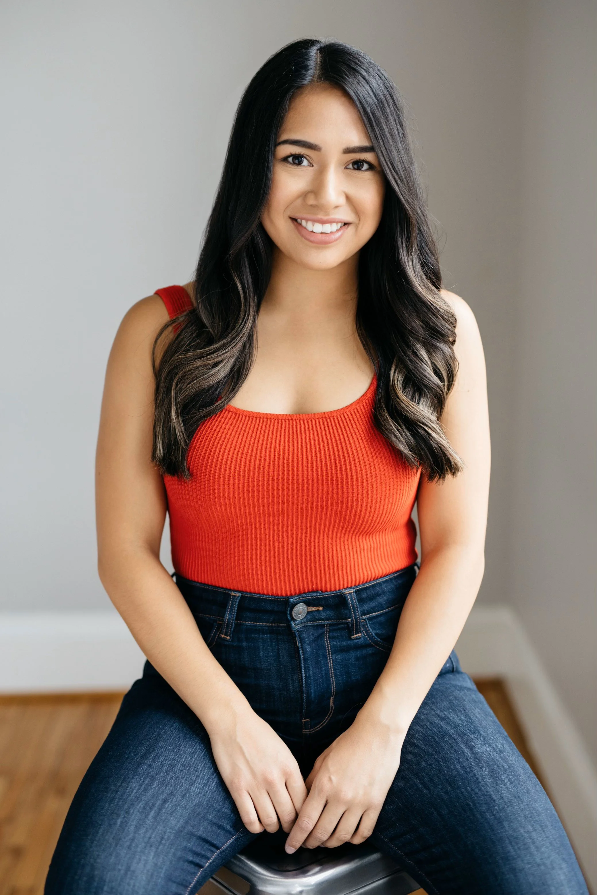 A young woman with long dark hair, smiling, wears a red sleeveless top and dark blue jeans, sitting on a stool indoors with a neutral background.