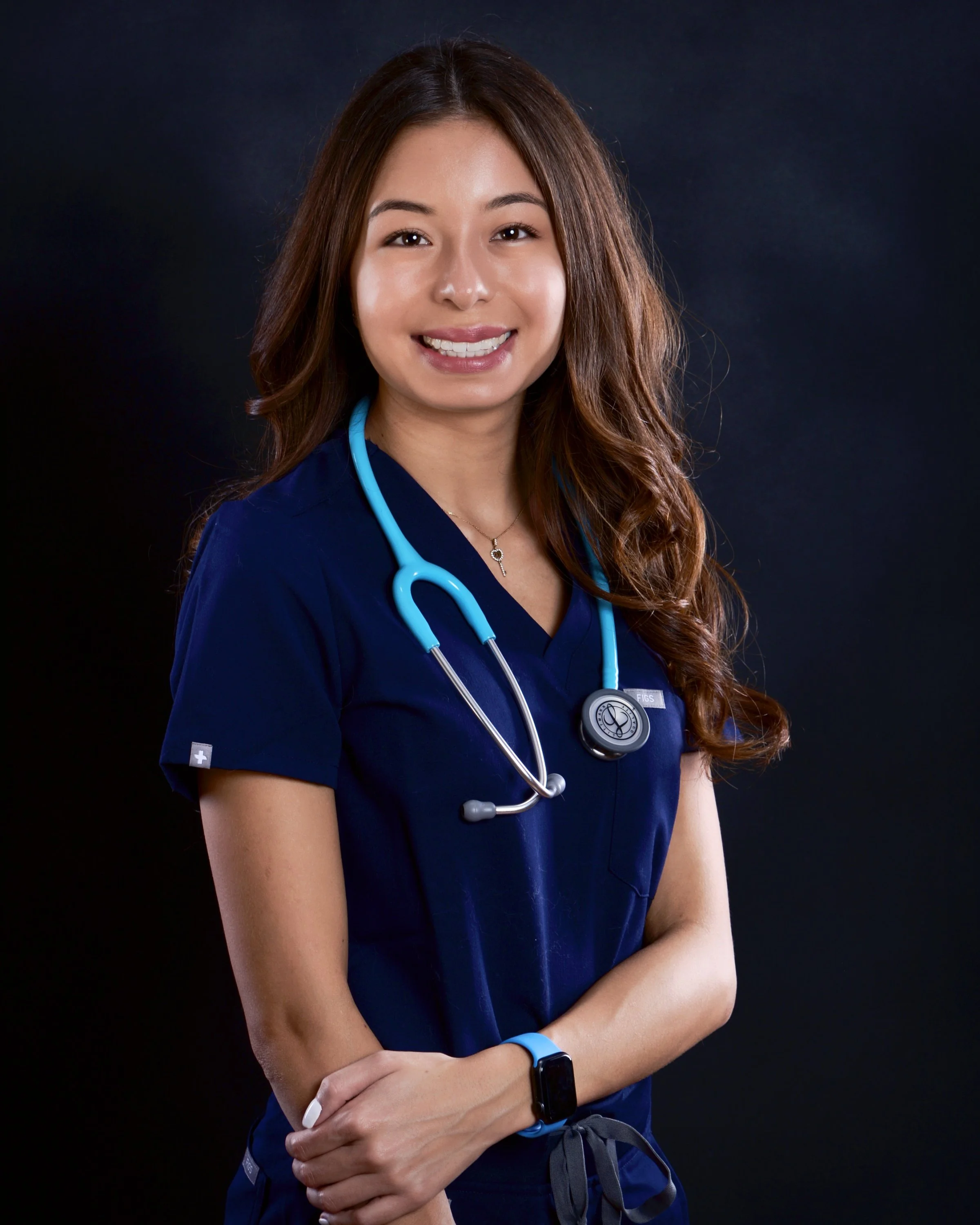A young woman in dark blue medical scrubs with a stethoscope around her neck, smiling at the camera against a dark background.
