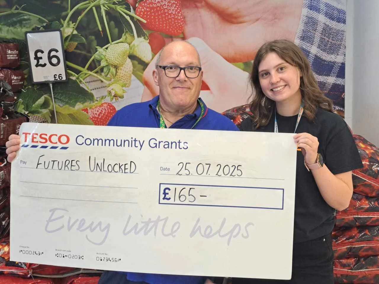 Two people hold a Tesco Community Grants cheque for £165, dated 25.07.2025, in front of a fruit display.