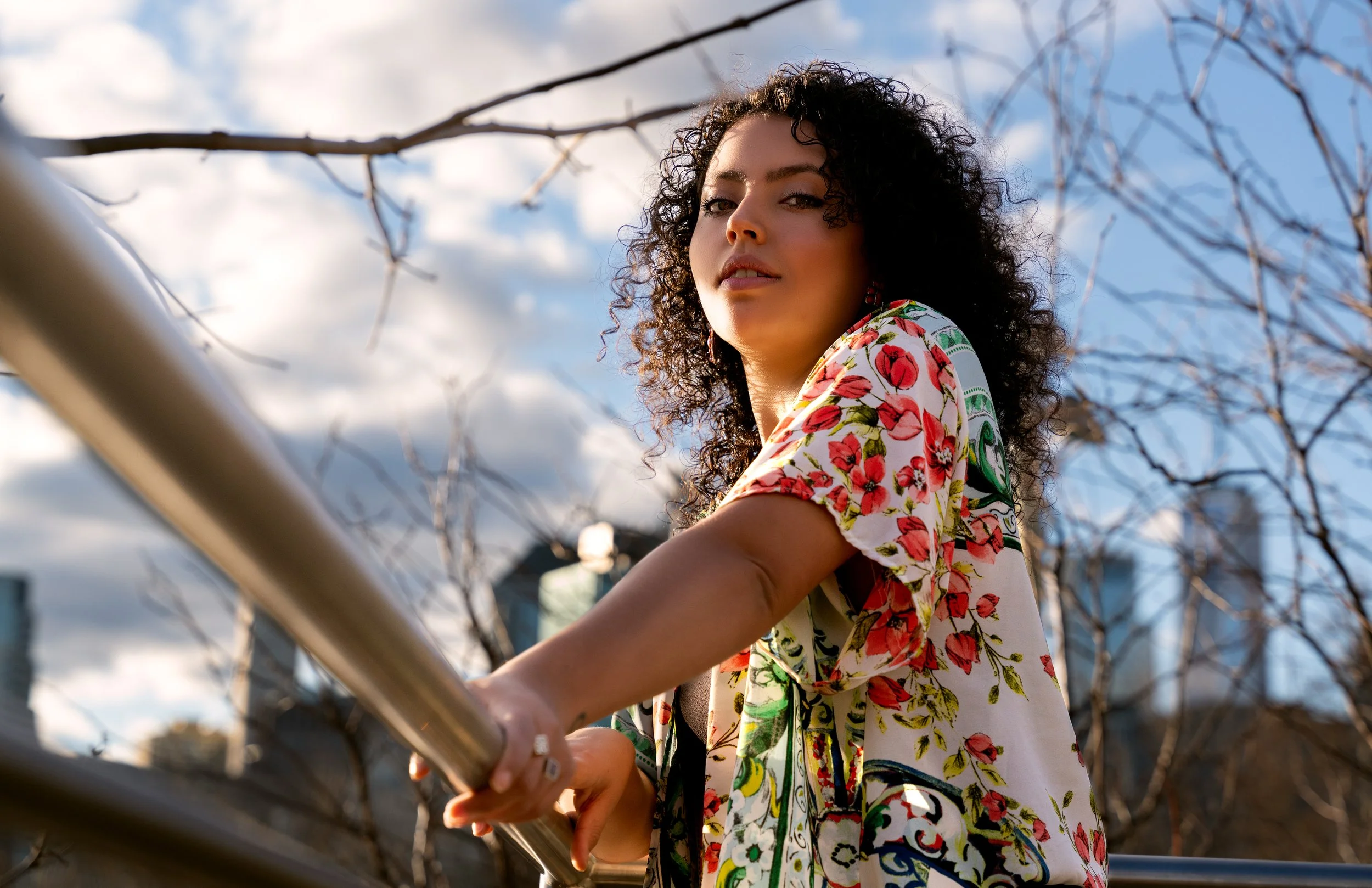 Model with curly hair leaning on metal railing, photographed from a low angle with city skyline and branches in background