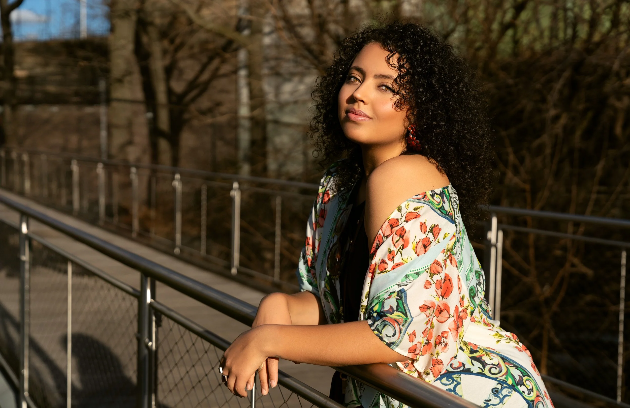Model in floral kimono standing by a walkway railing, captured in golden hour light with trees behind