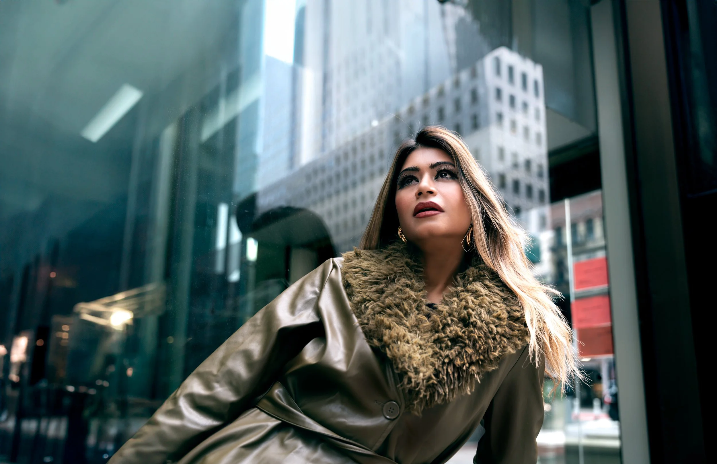 Editorial fashion portrait of Yesmin in a fur coat leaning against glass with city reflections in Manhattan