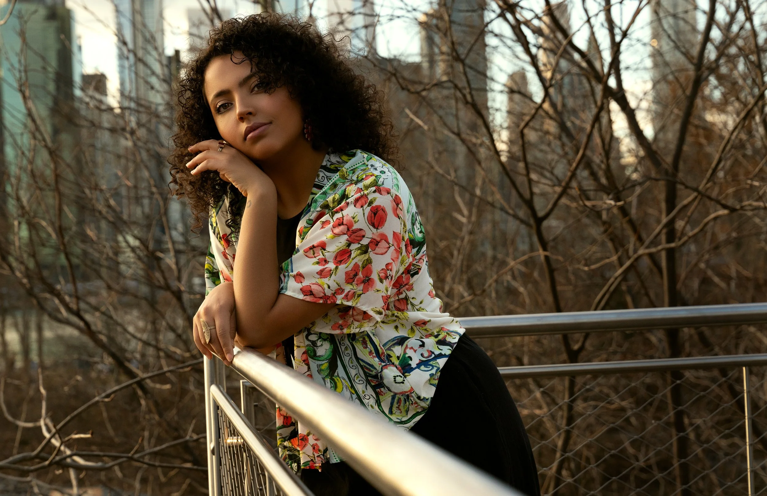 Editorial portrait of curly-haired model leaning on railing with Manhattan skyline in soft evening light