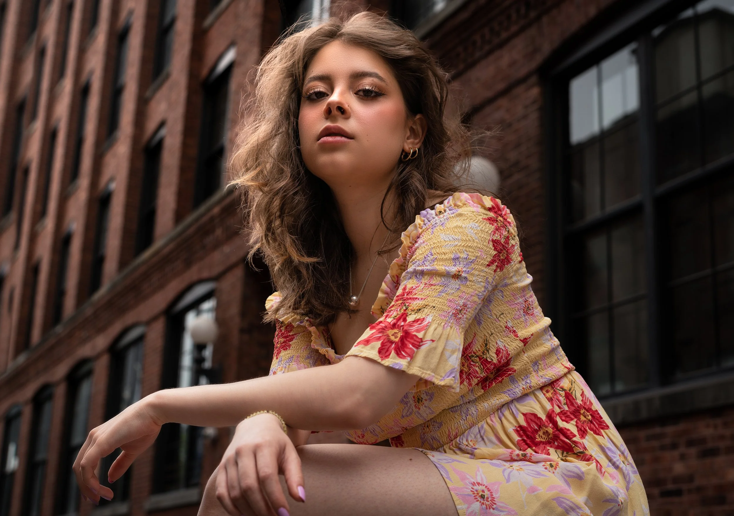 Editorial fashion portrait of Julianna sitting outdoors in a floral dress with a brick building background