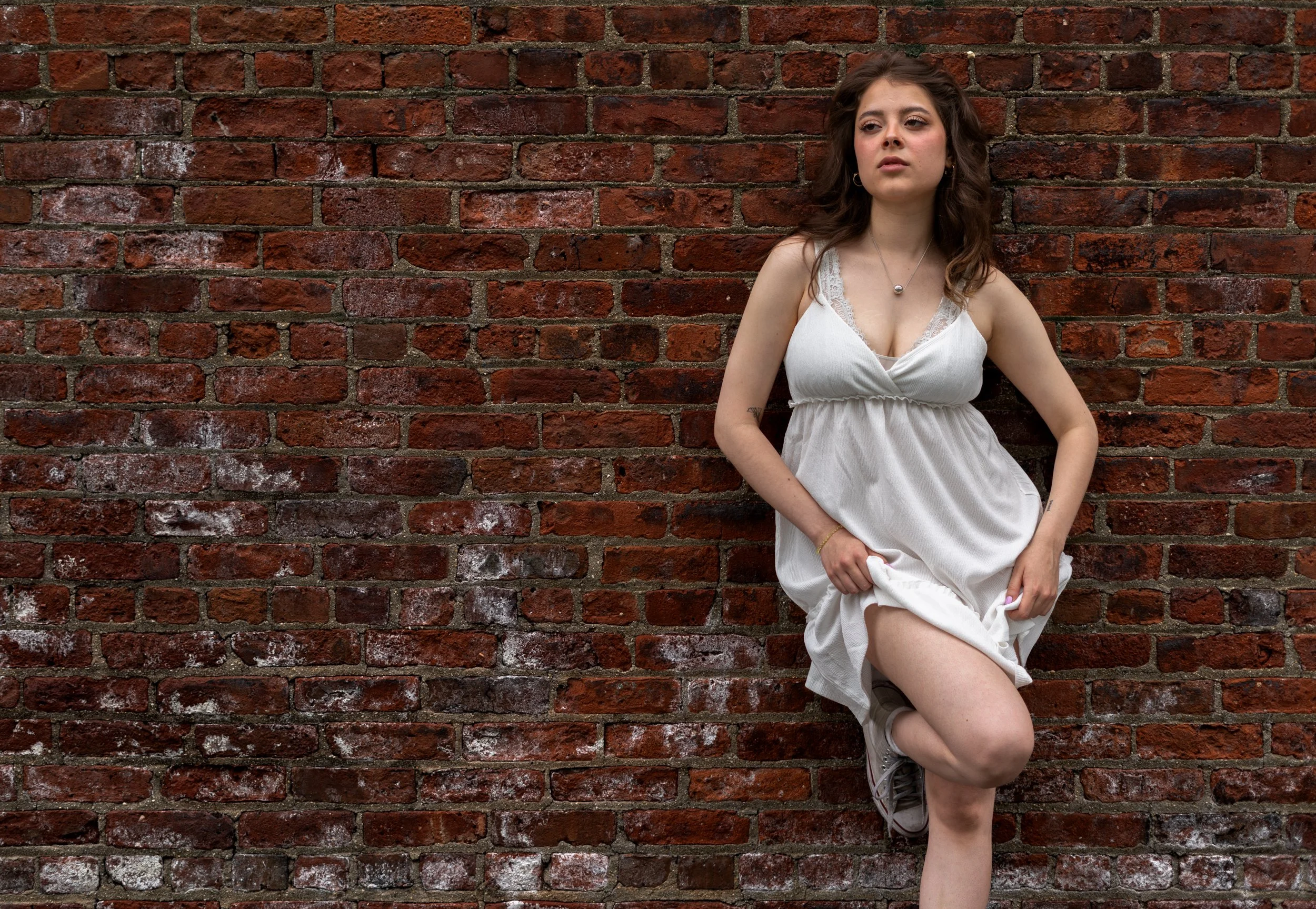 Full-body editorial portrait of Julianna in a white dress posing against a textured brick wall in NYC