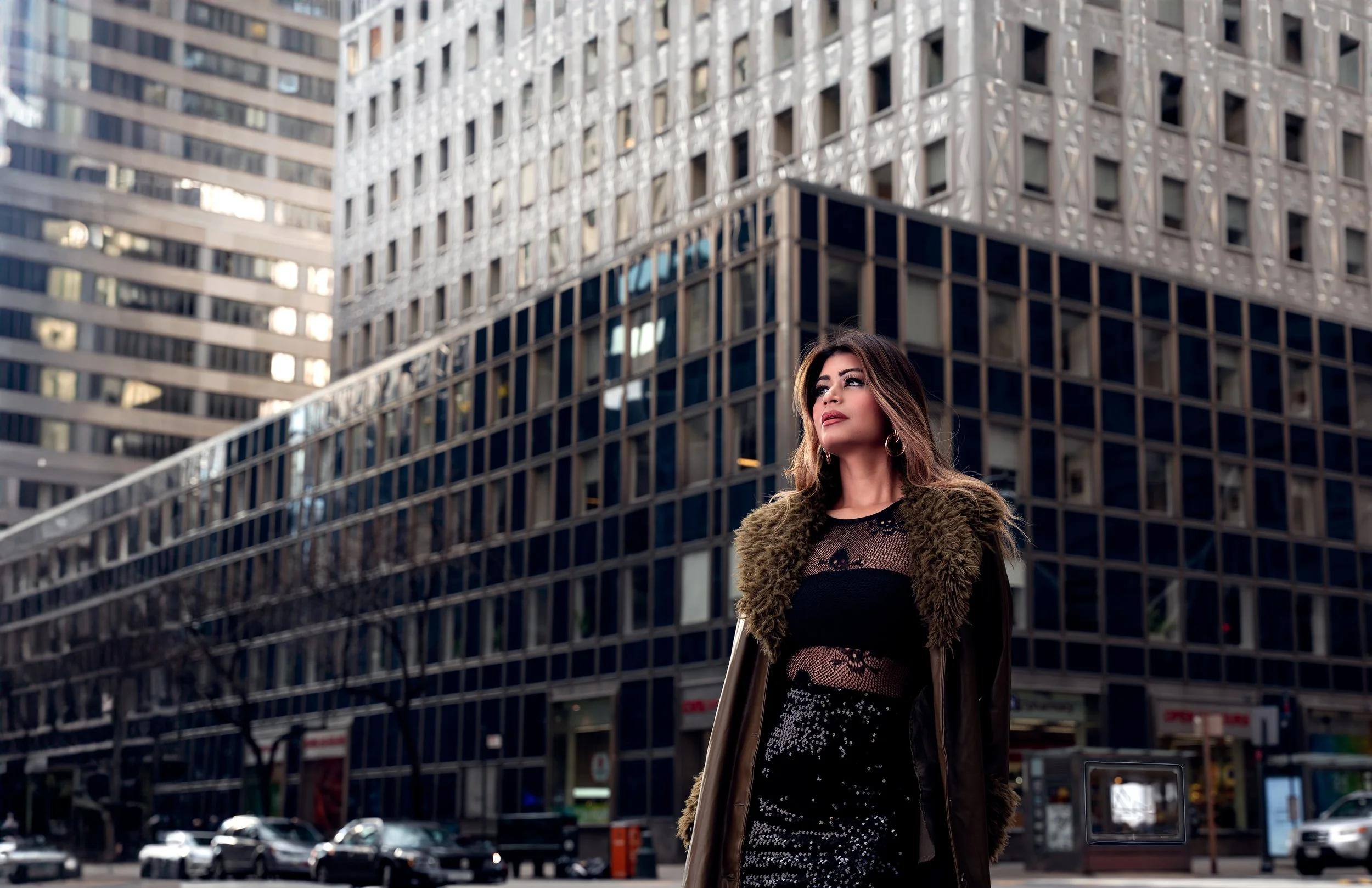 Wide editorial portrait of Yesmin in a fur coat standing in front of modern buildings in NYC