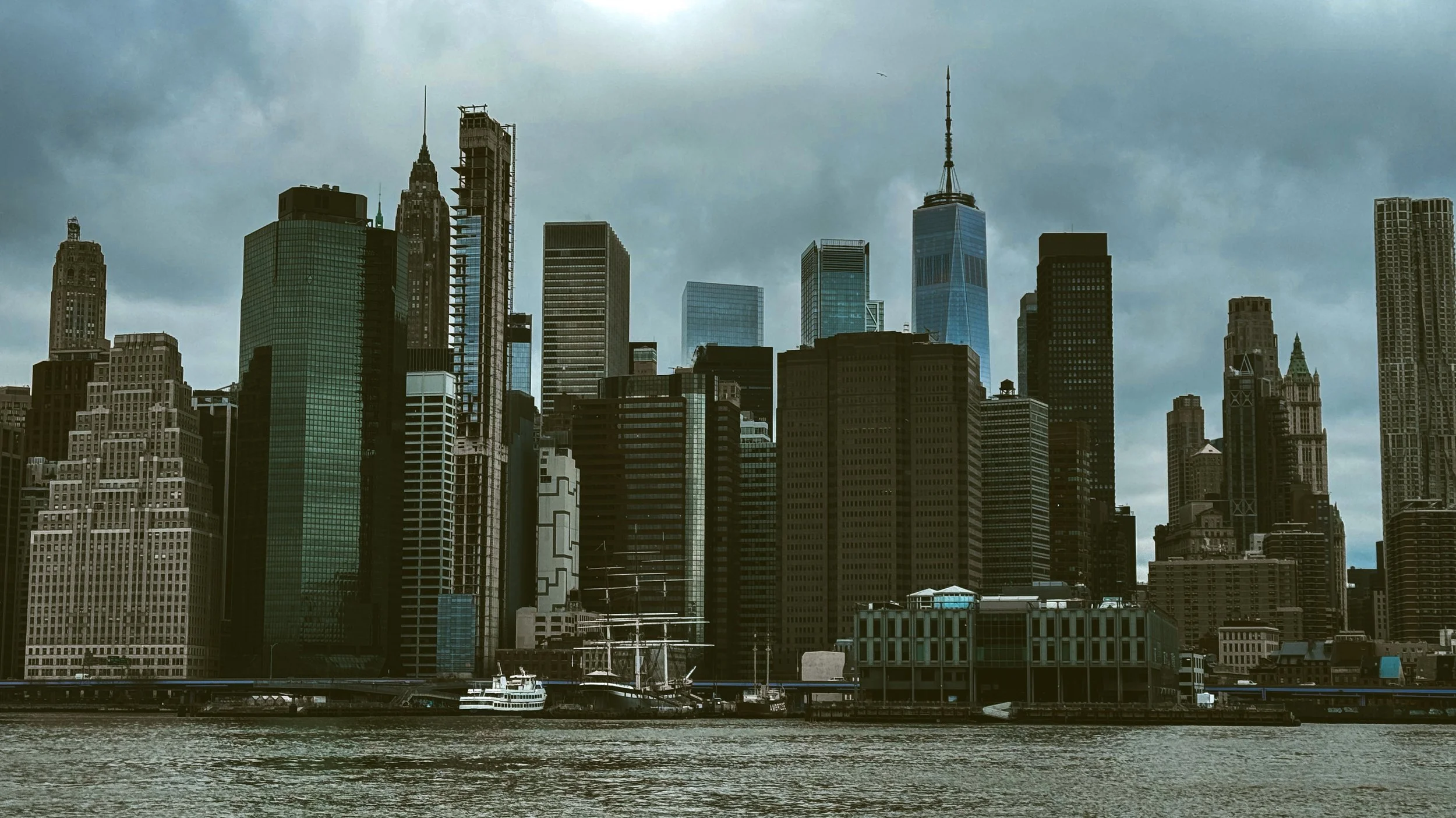 Lower Manhattan skyline in New York City featuring a mix of historic and modern skyscrapers along the waterfront under dramatic sky