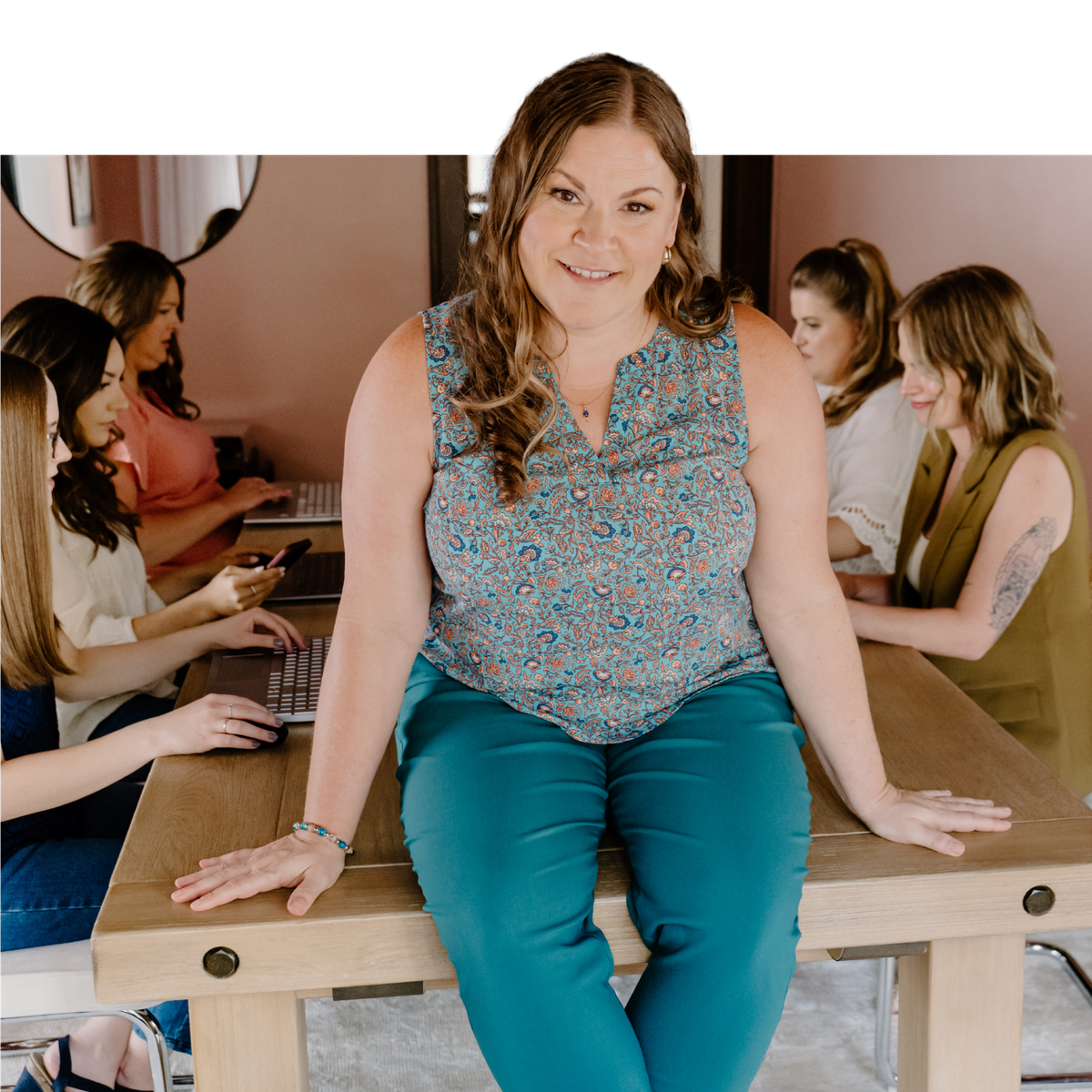 aTiffany Bastian sitting on the office table while her team works on laptops behind her