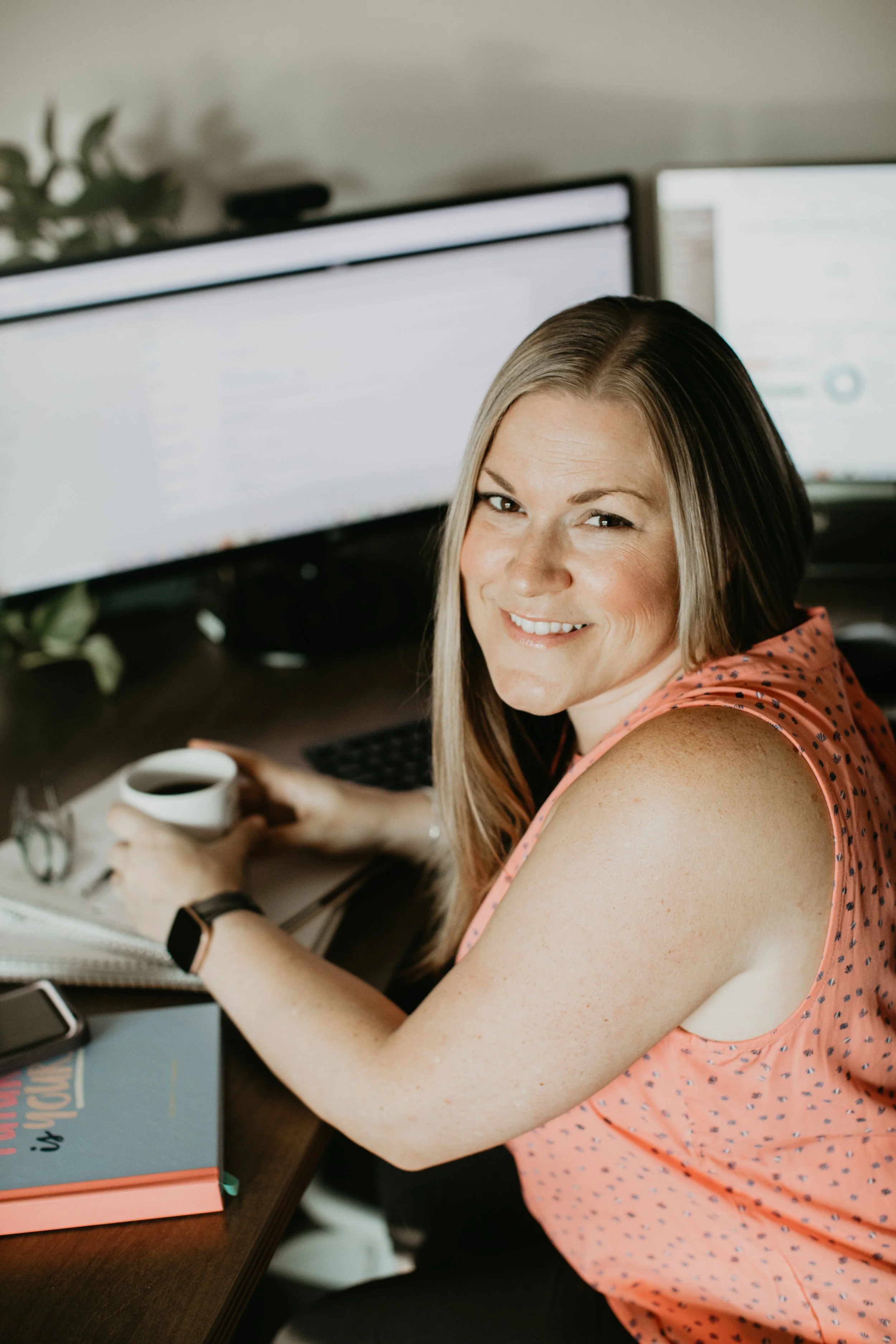 Tiffany Bastian smiling at her desk with a coffee mug, in front of two monitors