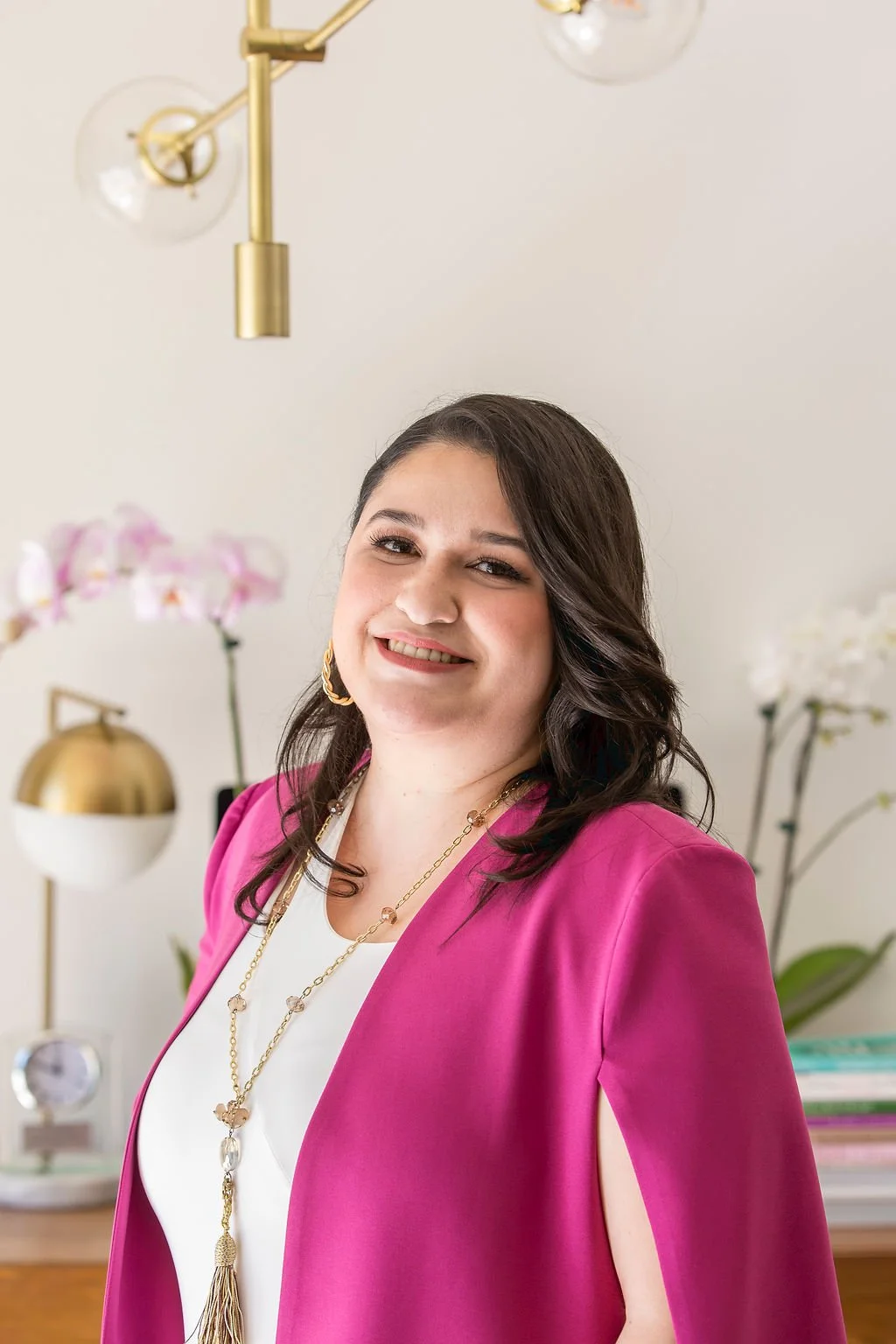 In this image we can see Mishka Kimball LMTF, smiling with her hair down, wearing a pink blazer and a white center, in the background we can see some beautiful orchids.