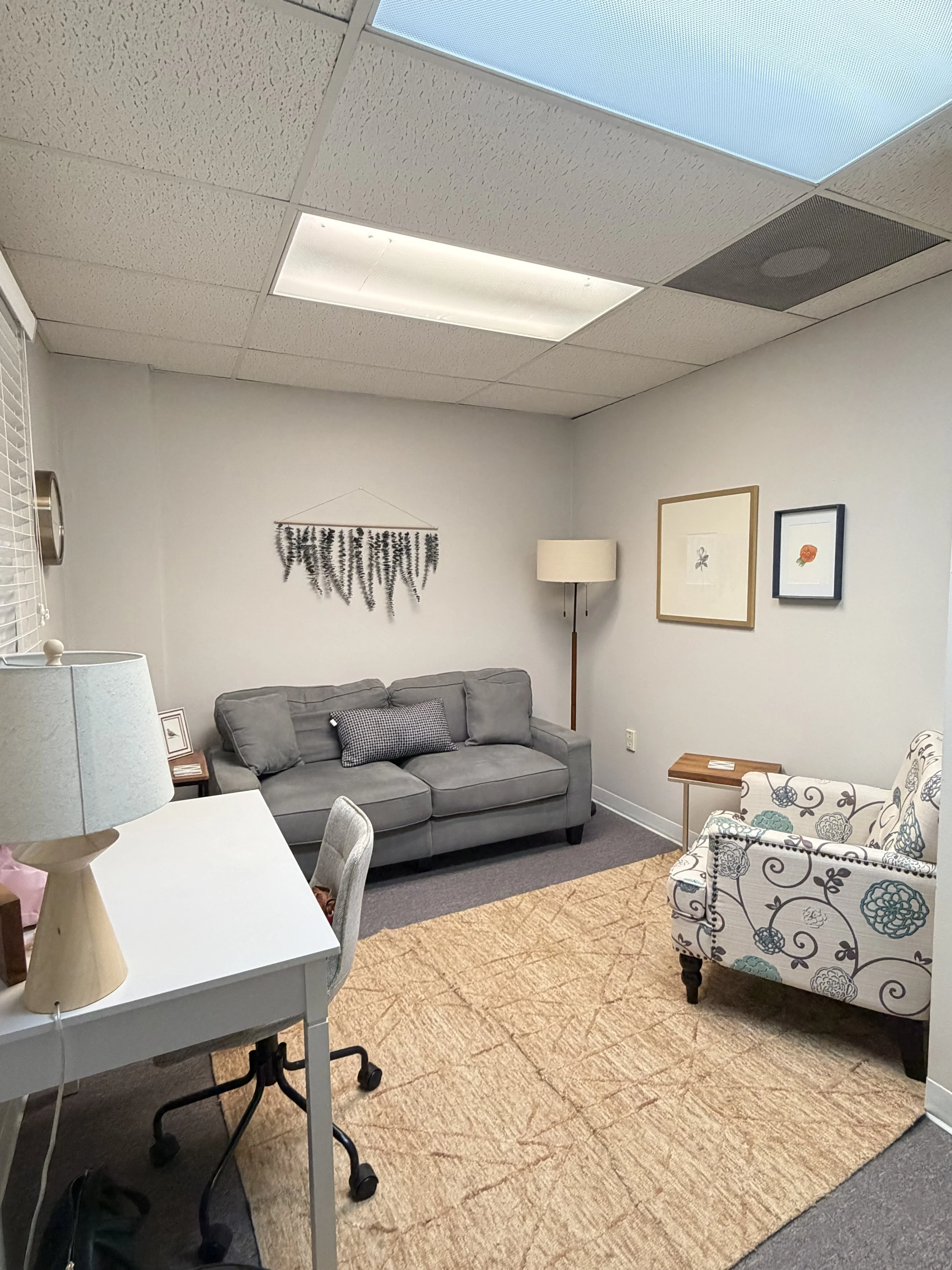 A cozy office with light-colored walls, a gray sofa, a floral armchair, framed artwork, a wooden side table, and a beige area rug. This is a therapy office where clients can feel comfortable.