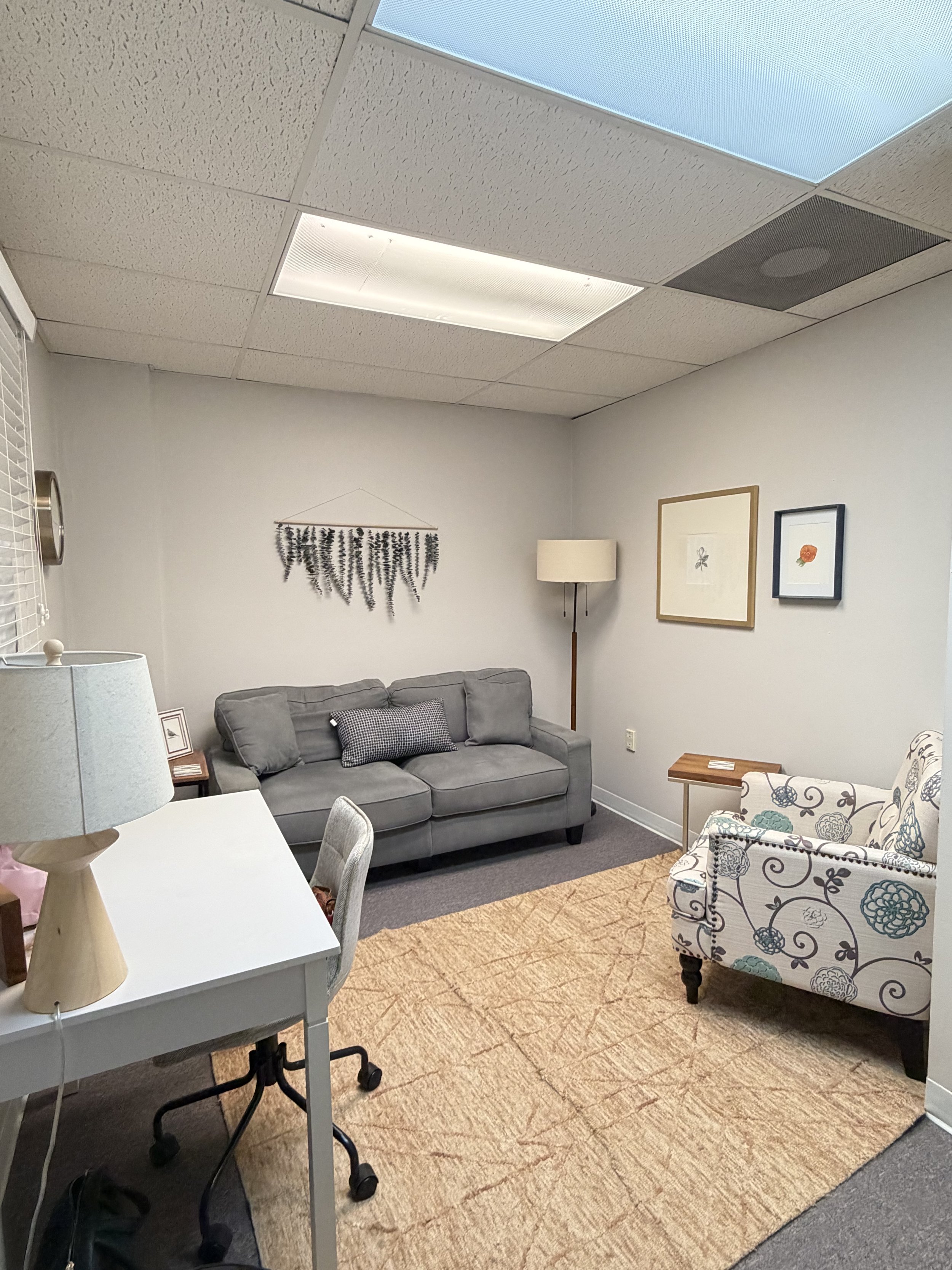 A cozy office with light-colored walls, a gray sofa, a floral armchair, framed artwork, a wooden side table, and a beige area rug. This is a therapy office where clients can feel comfortable.