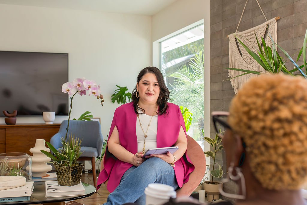 Mishka Kimball, LMFT, in a pink jacket sitting on a chair holding a tablet, in a cozy, well-decorated living room. There are plants, a coffee table with books and decorative items, and a large window with natural light.
