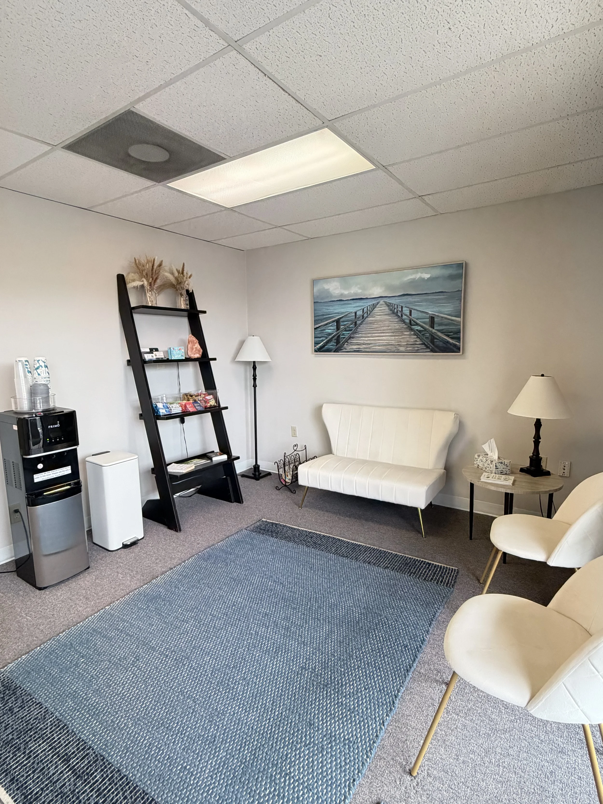 An indoor waiting room with a blue woven rug, white chairs, a white loveseat, a black ladder-style shelf with decorative items, a white floor lamp, a framed seascape painting, a side table with a lamp and tissue box, & a water dispenser.
