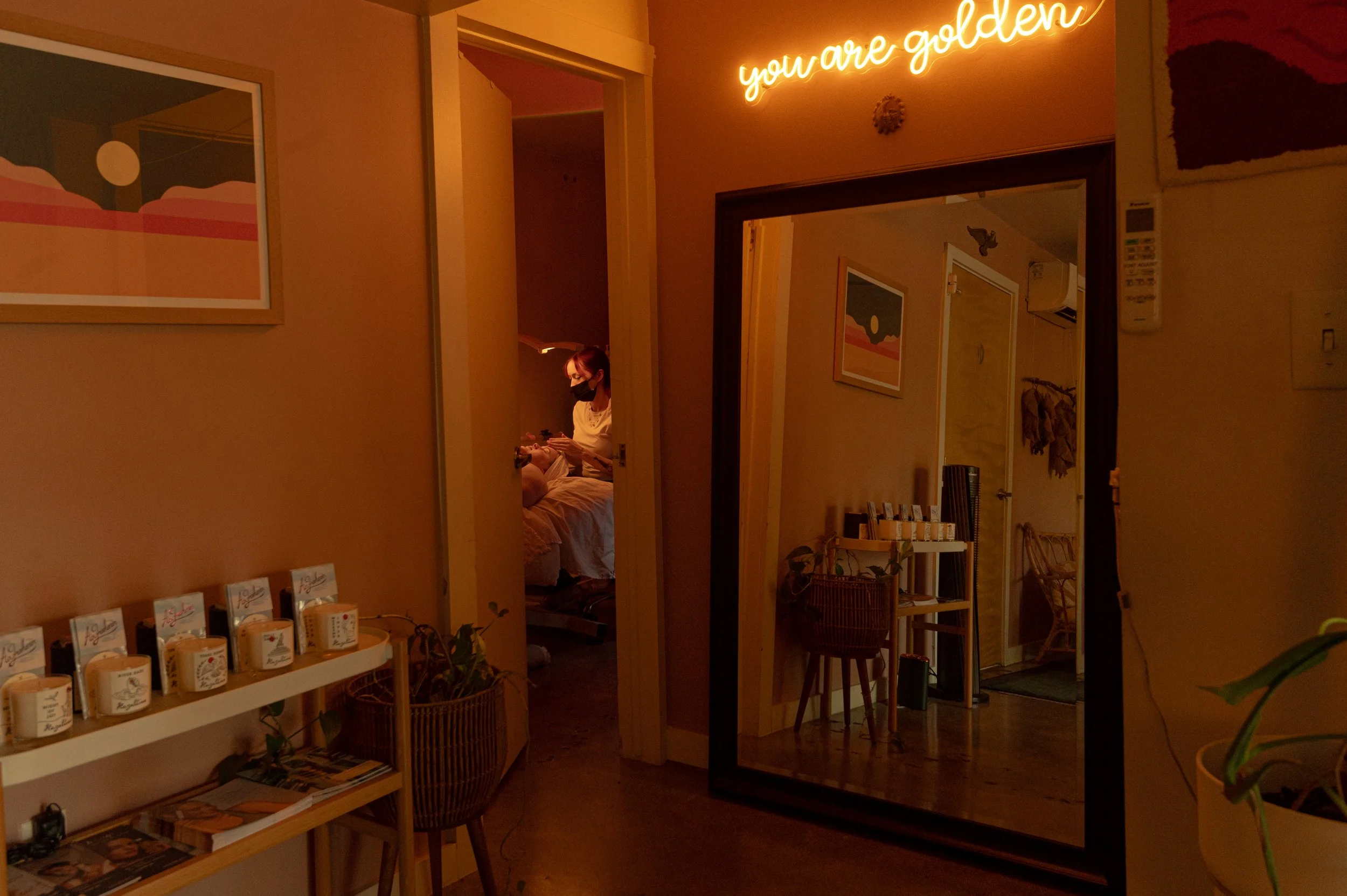 Interior of a pharmacy or skincare store with wooden shelves holding skincare products, plants, and decorative items, and a small wooden display table below. There are indoor plants on both sides.