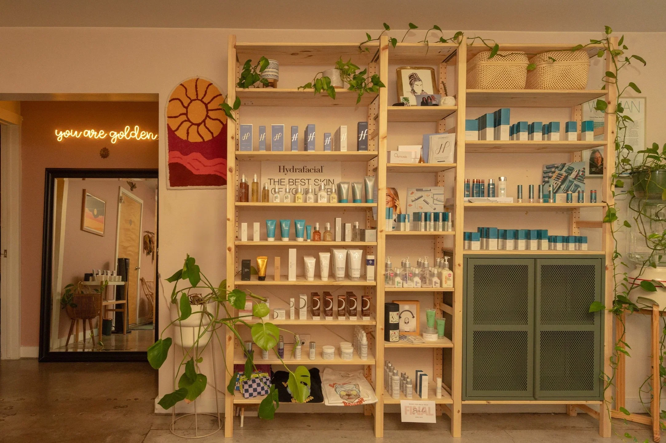 Interior view of a cozy room with pink walls, decorated with plants, a rainbow-shaped rug, and various furniture including chairs, shelves, and a water dispenser. The room features wooden accents and a partially open door leading to a tiled area.