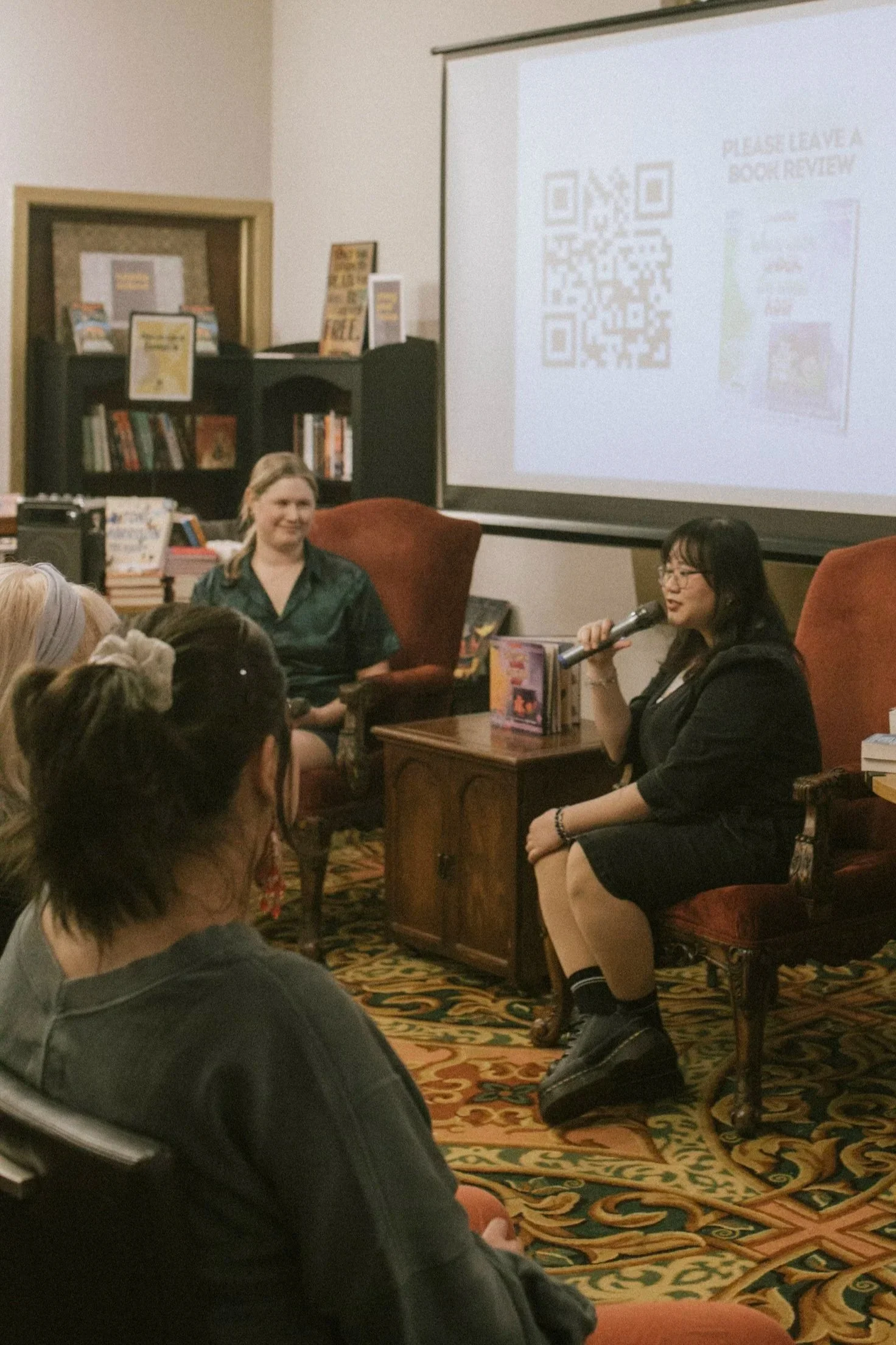 A woman with glasses holding a microphone and speaking in front of an audience at a book review event, with a bookshelf and a large screen displaying a QR code and event information in the background.