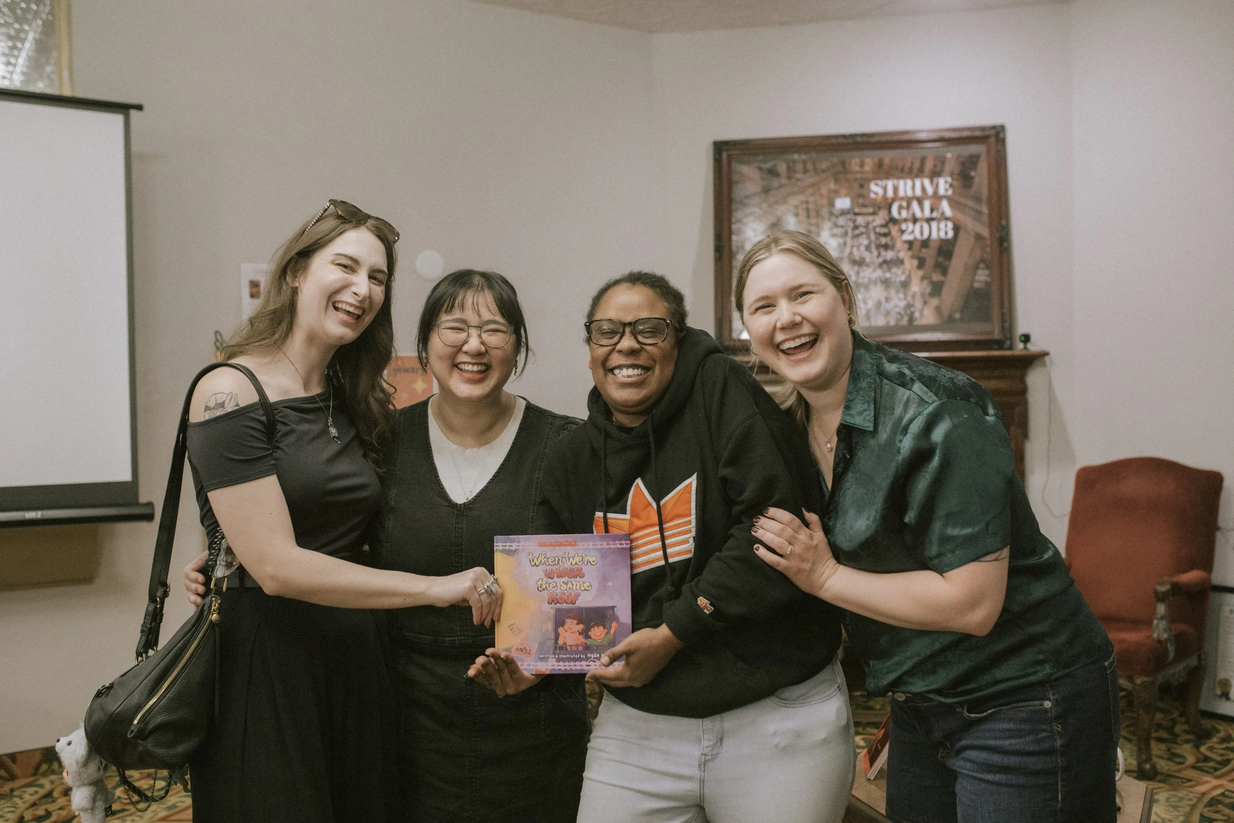 Four women are standing together indoors, smiling and hugging. They are holding a colorful children's book titled 'When We're Under the Same Roof.' The background features a framed poster for the 2018 Strive Gala and a television screen.
