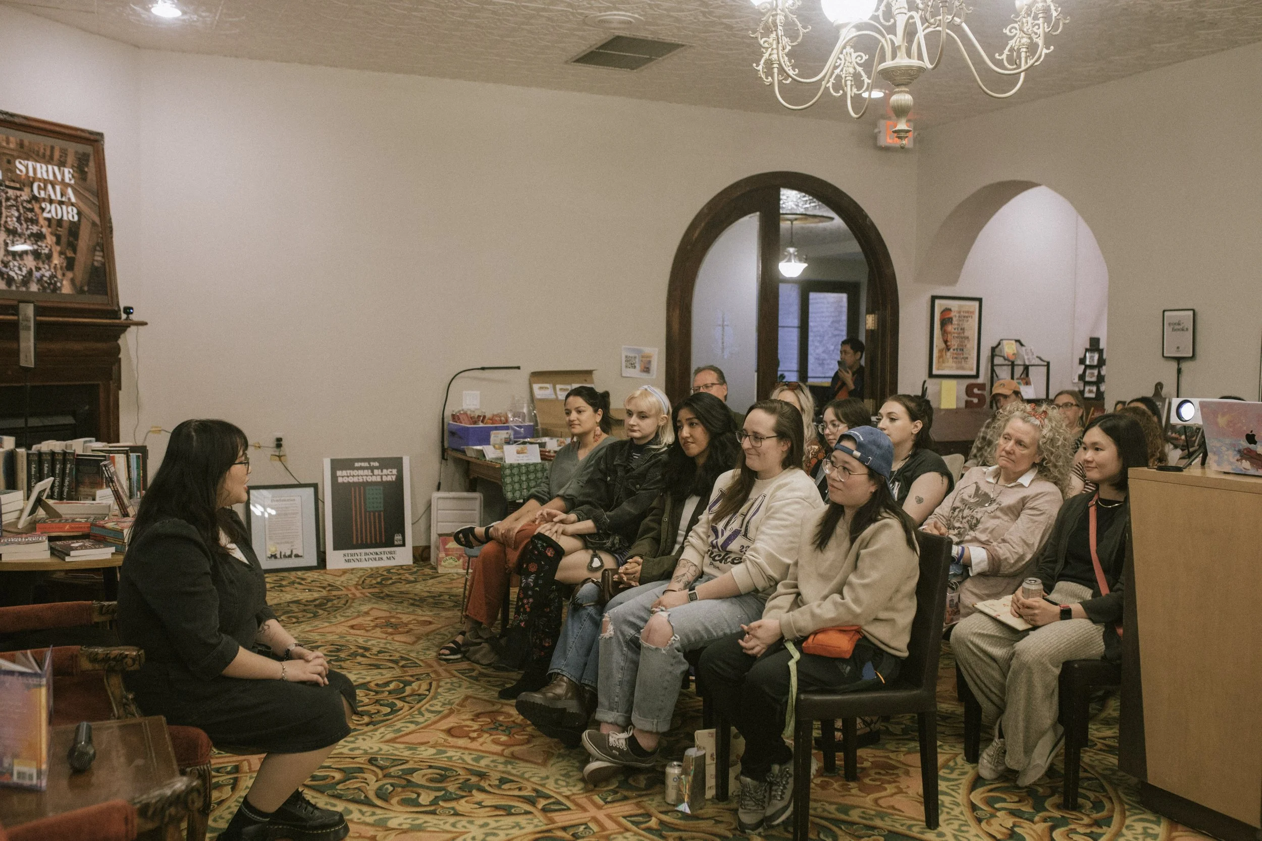 A woman speaking to an audience seated in a room with bookshelves, framed pictures, and a chandelier