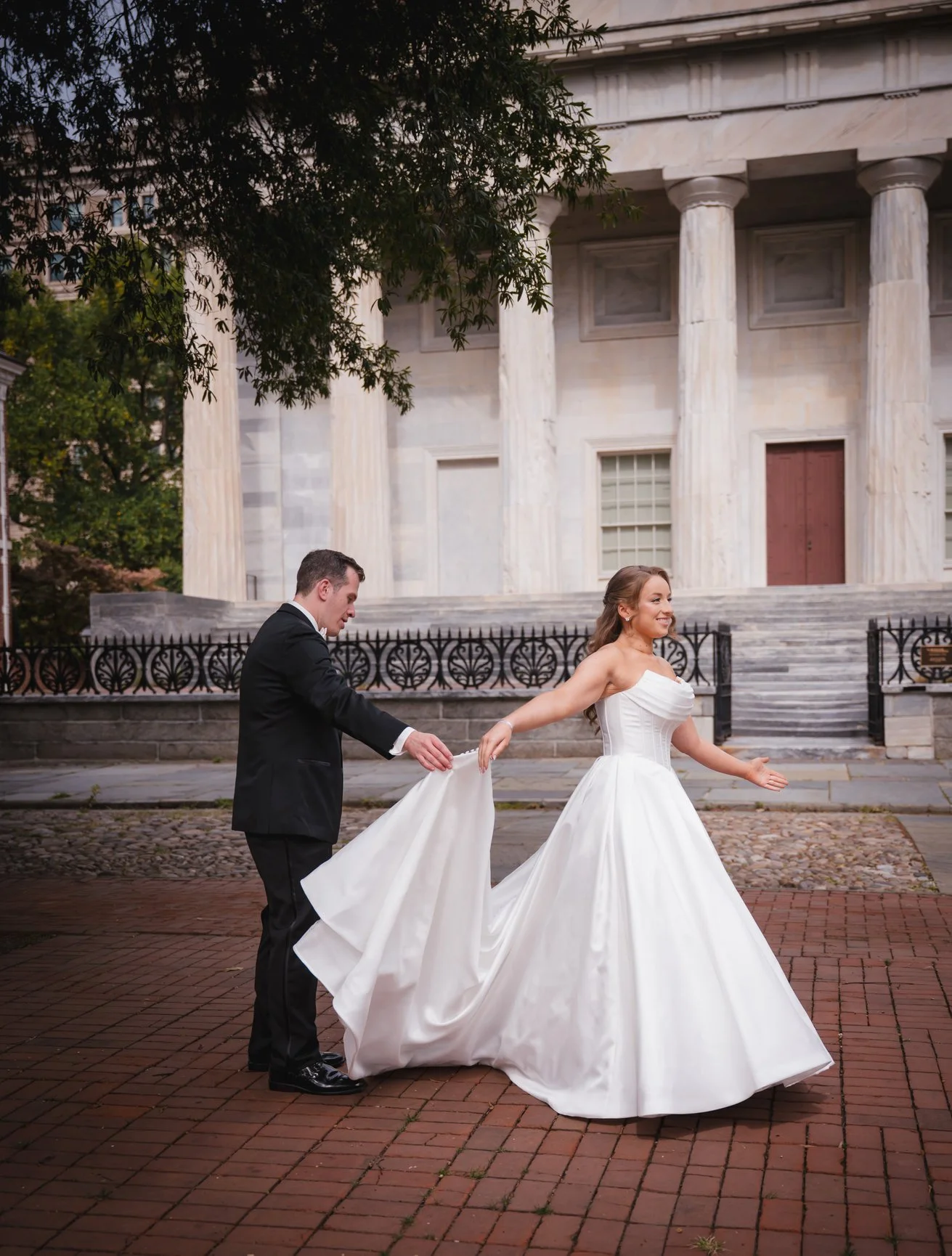 A bride and groom holding hands and dancing outside a historic building with large columns, cobblestone pavement, and tree branches in the foreground.