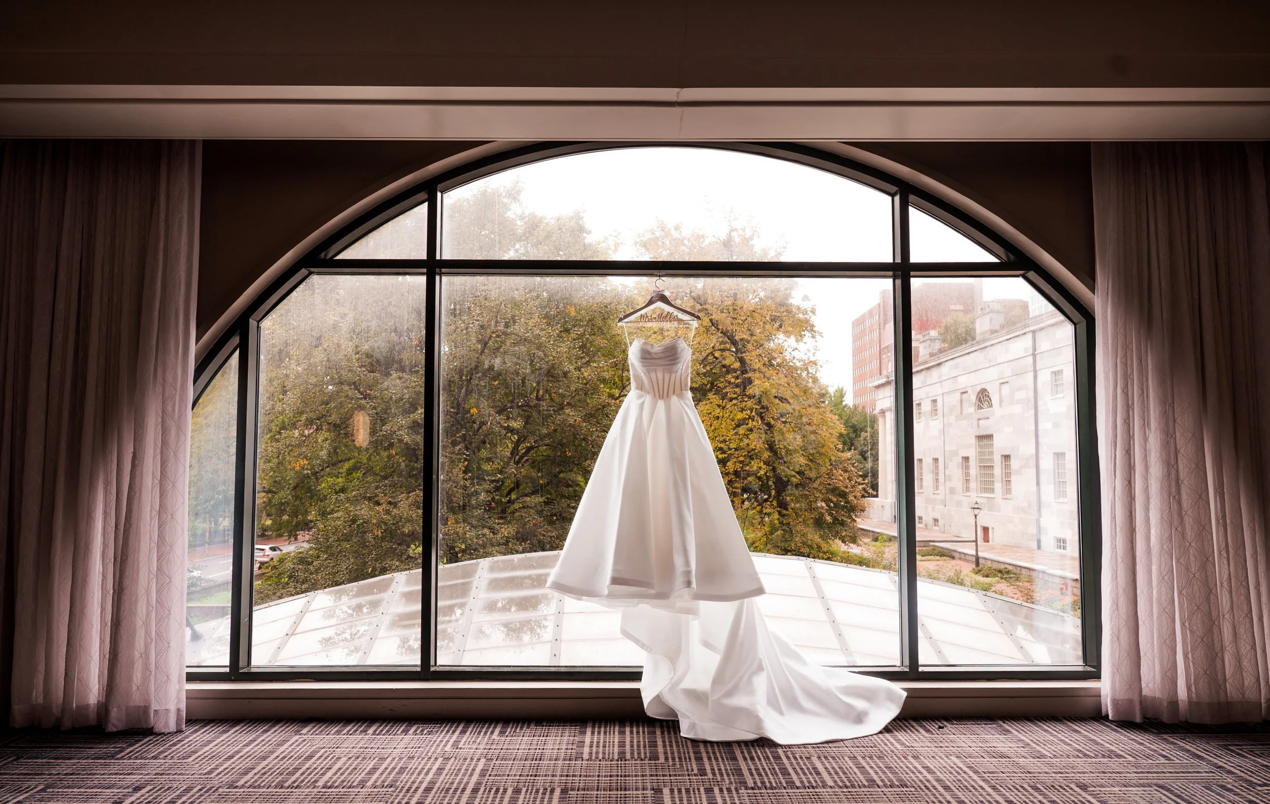 A white wedding dress hanging on a hanger in front of a large arched window overlooking trees and buildings.