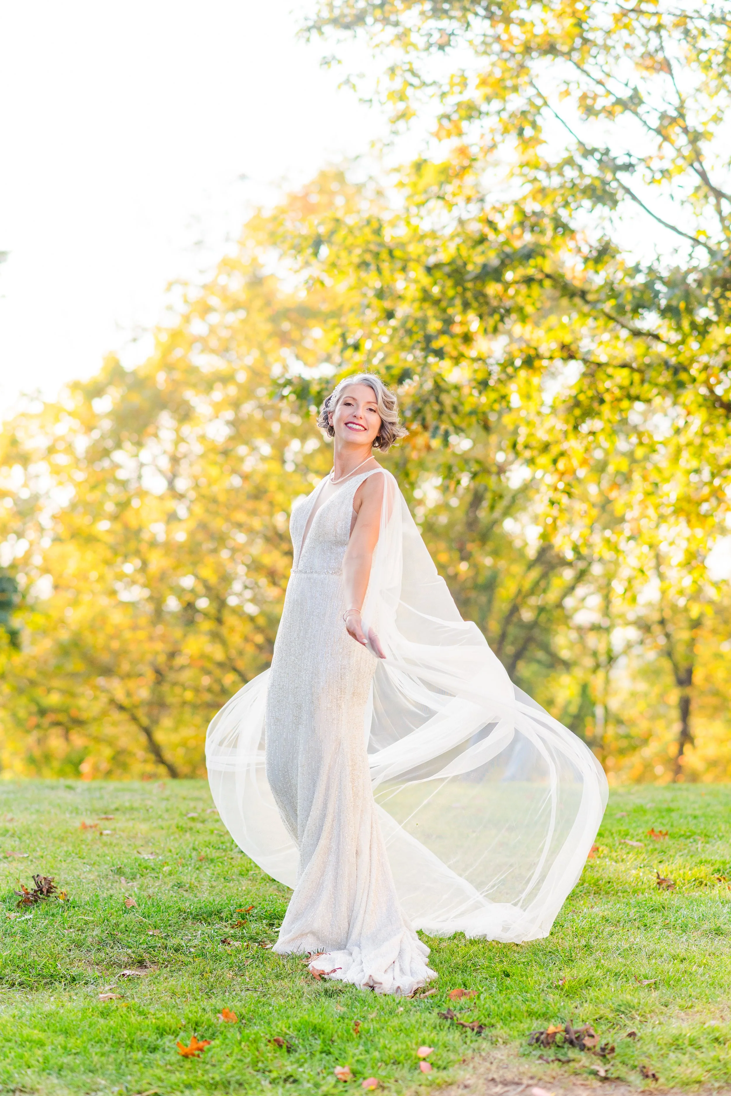 A smiling woman in a flowing white gown stands on grassy ground with autumn trees and sunlight in the background.