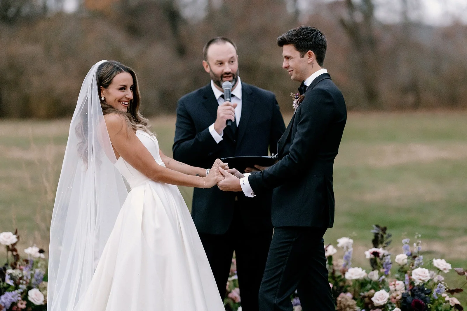 A bride and groom holding hands during an outdoor wedding ceremony with an officiant in the background holding a microphone, all smiling and dressed formally, with flowers around them on the grass field. strapless satin ballgown wona concept bridal d
