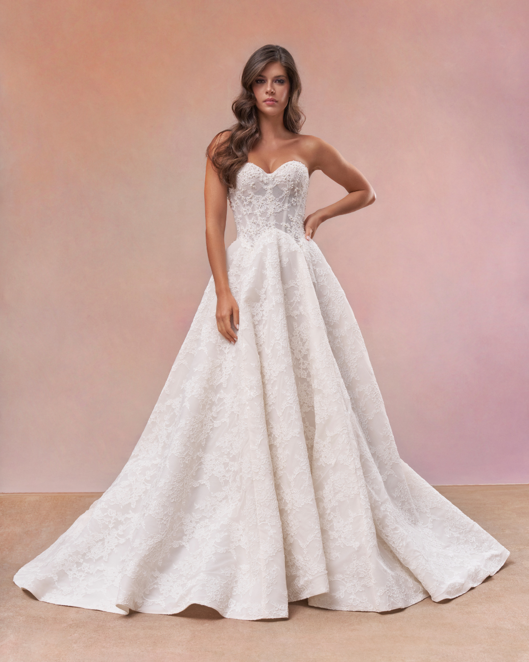 A woman wearing a strapless wedding gown with lace details, standing against a pastel pink and peach background. maria farbinni